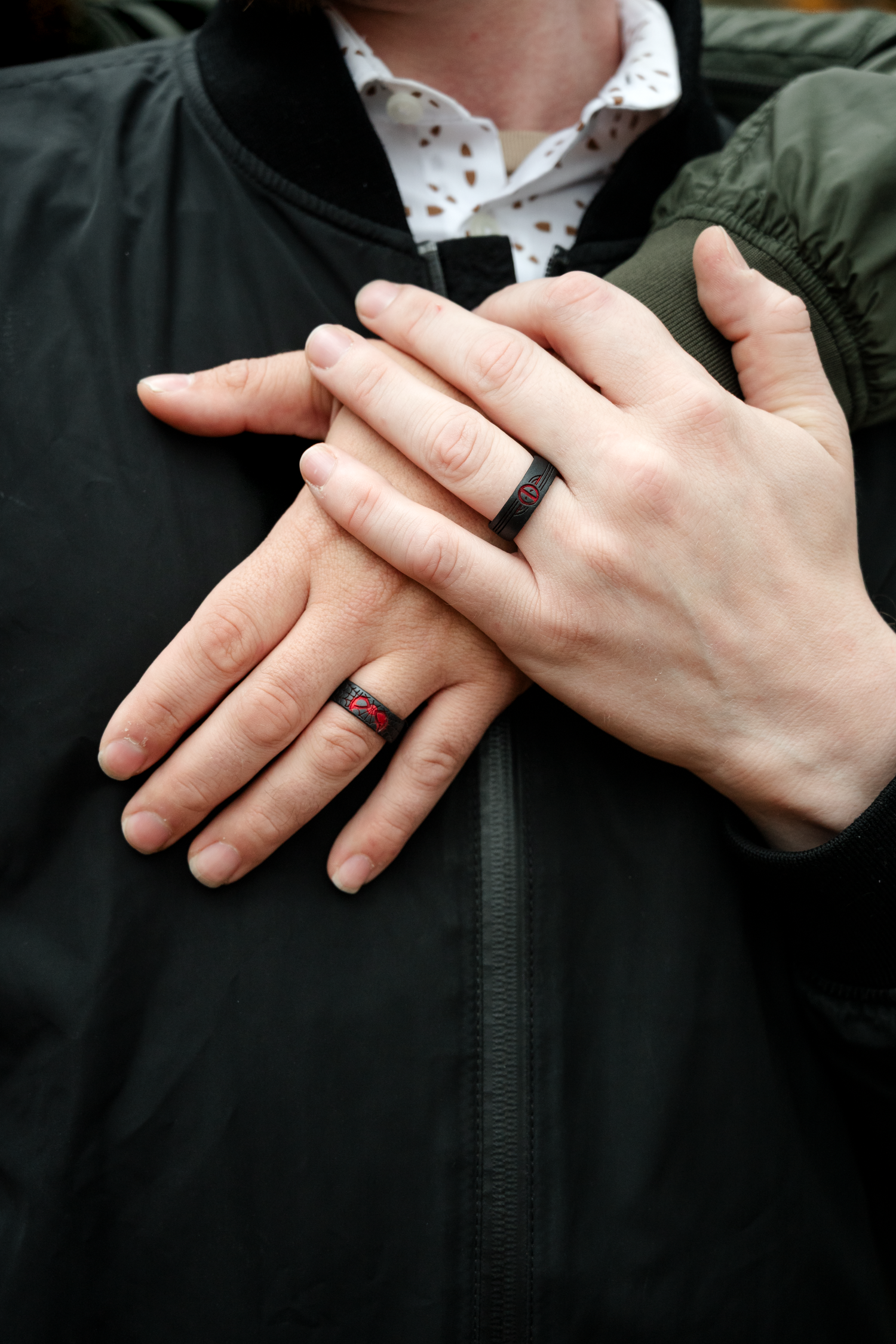 Two hands with matching black and red silicone wedding bands crossed over a dark jacket.