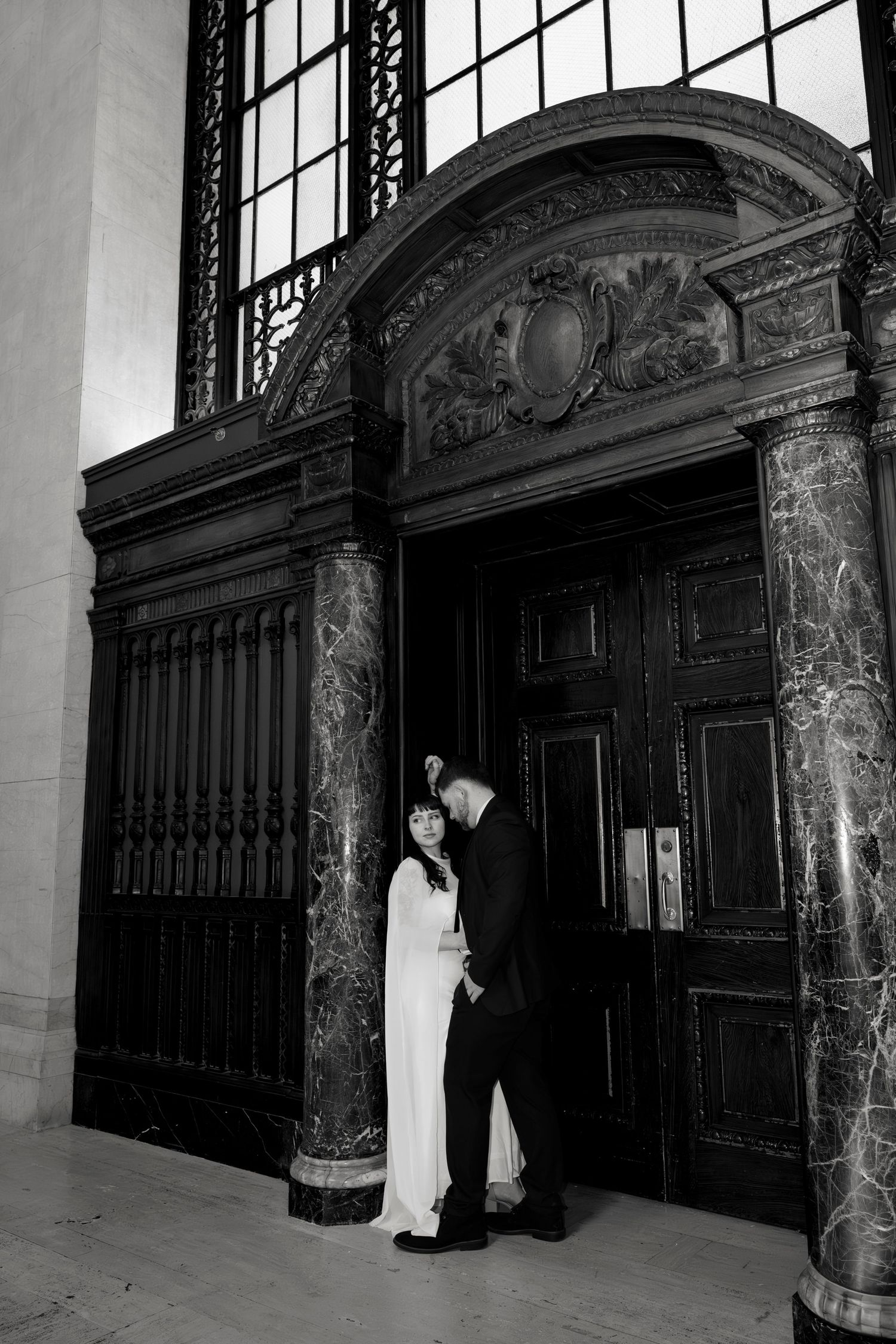 Bride and groom embrace by ornate wooden doors with marble columns in black and white photo.