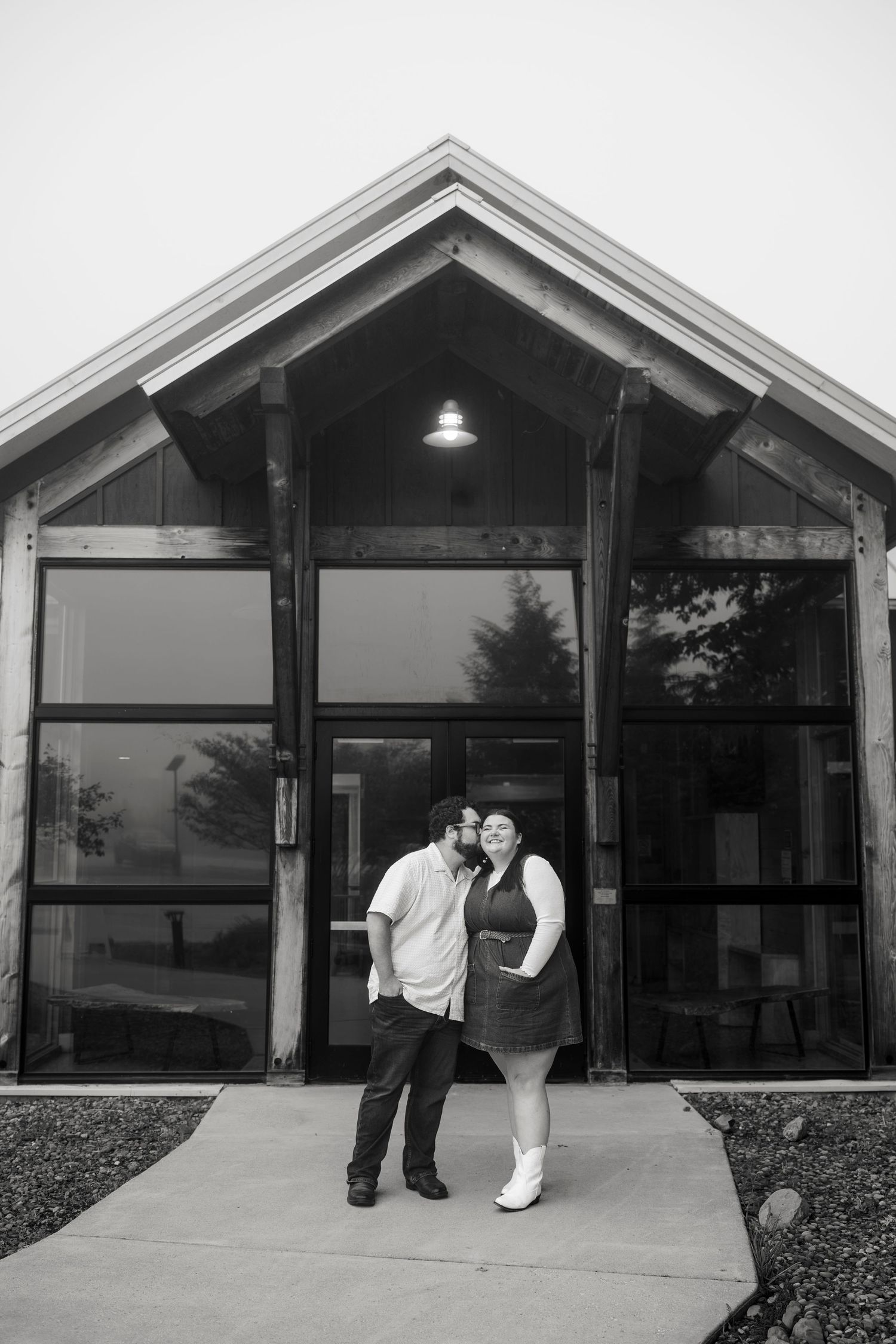 Couple poses romantically in front of a modern wood and glass building entrance in black and white photo.