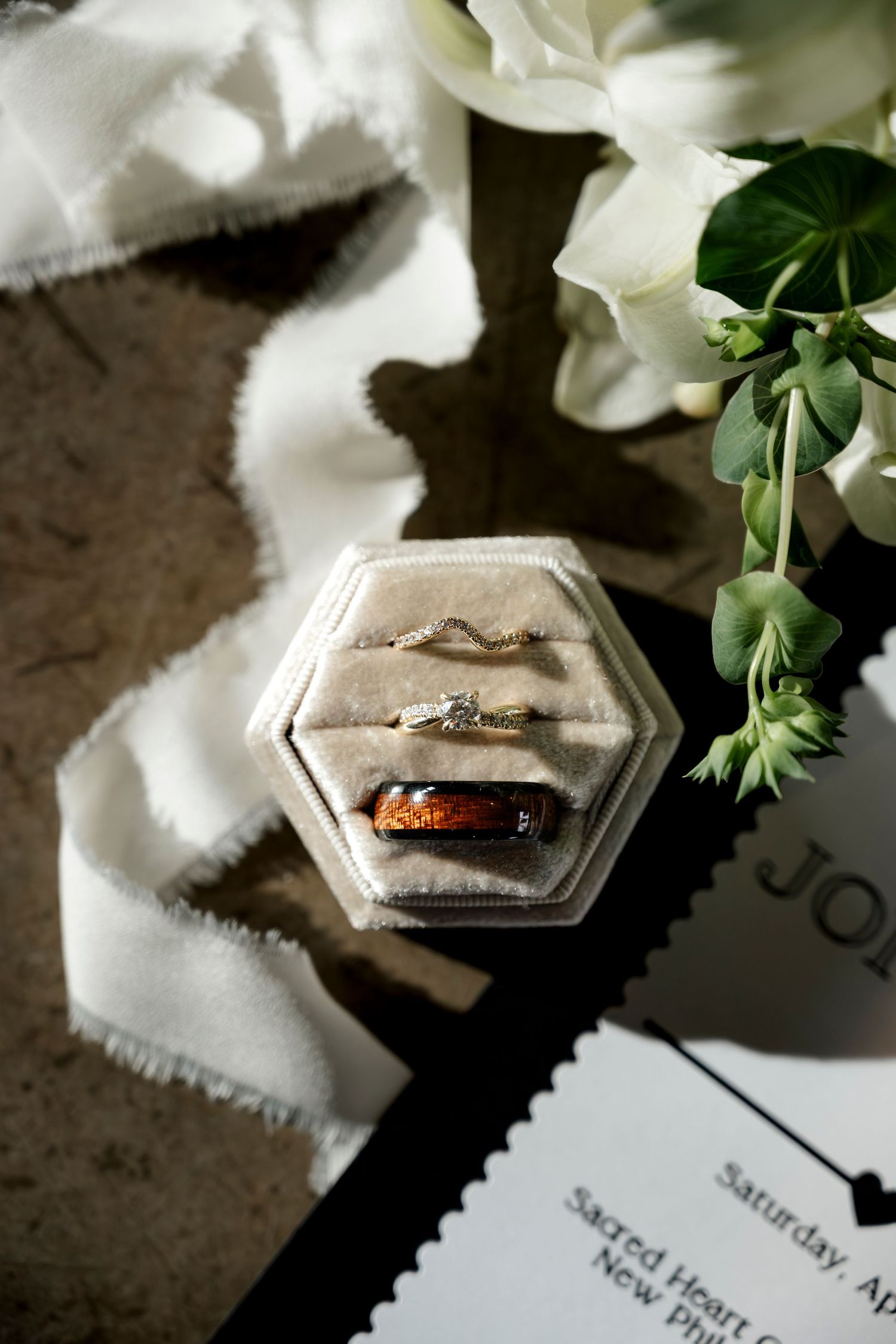 Wedding rings in a hexagonal velvet box beside white flowers and a wedding invitation on a marble surface.