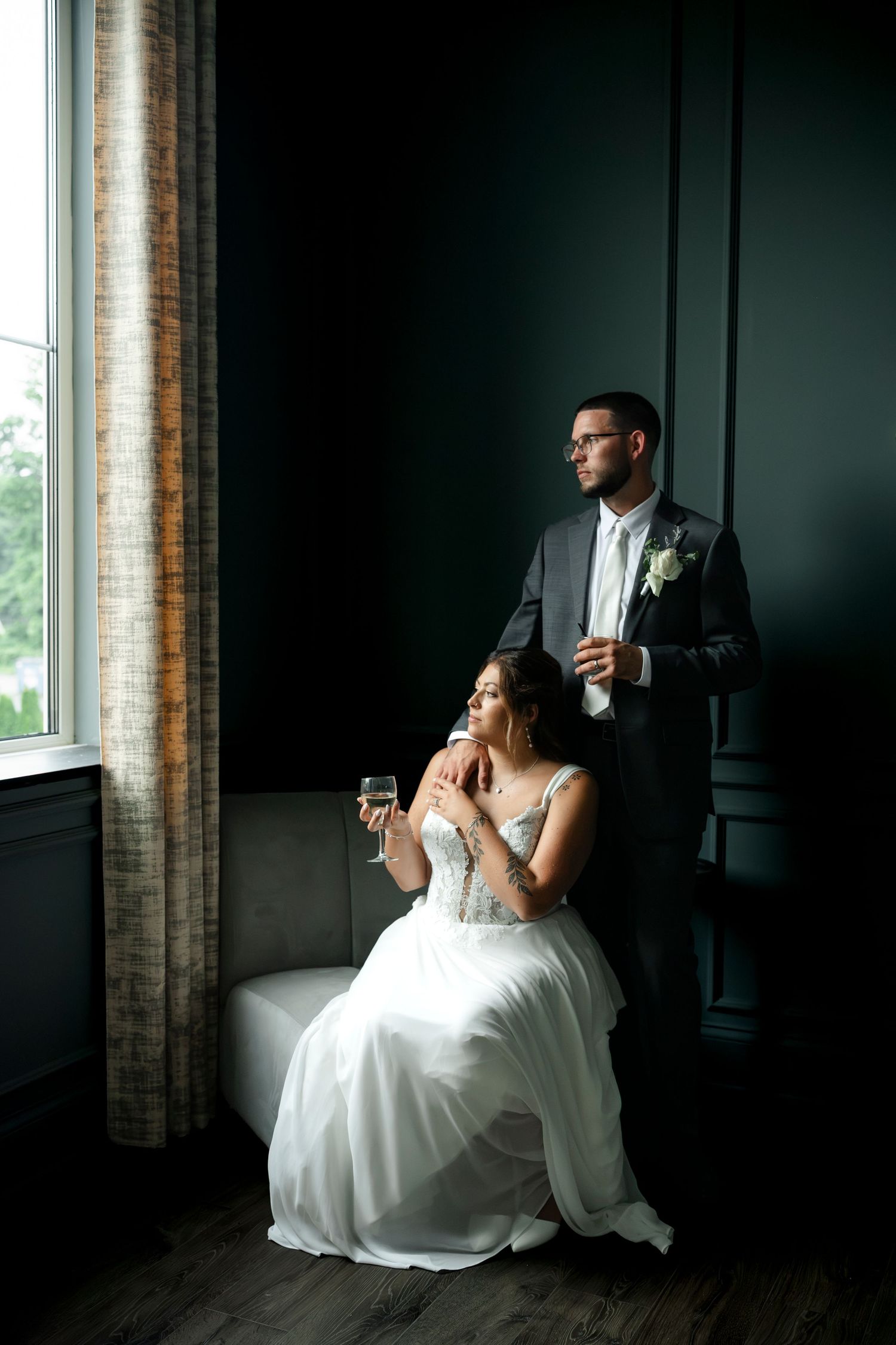 Bride in white gown sits holding champagne while groom in gray suit stands beside her near a window.