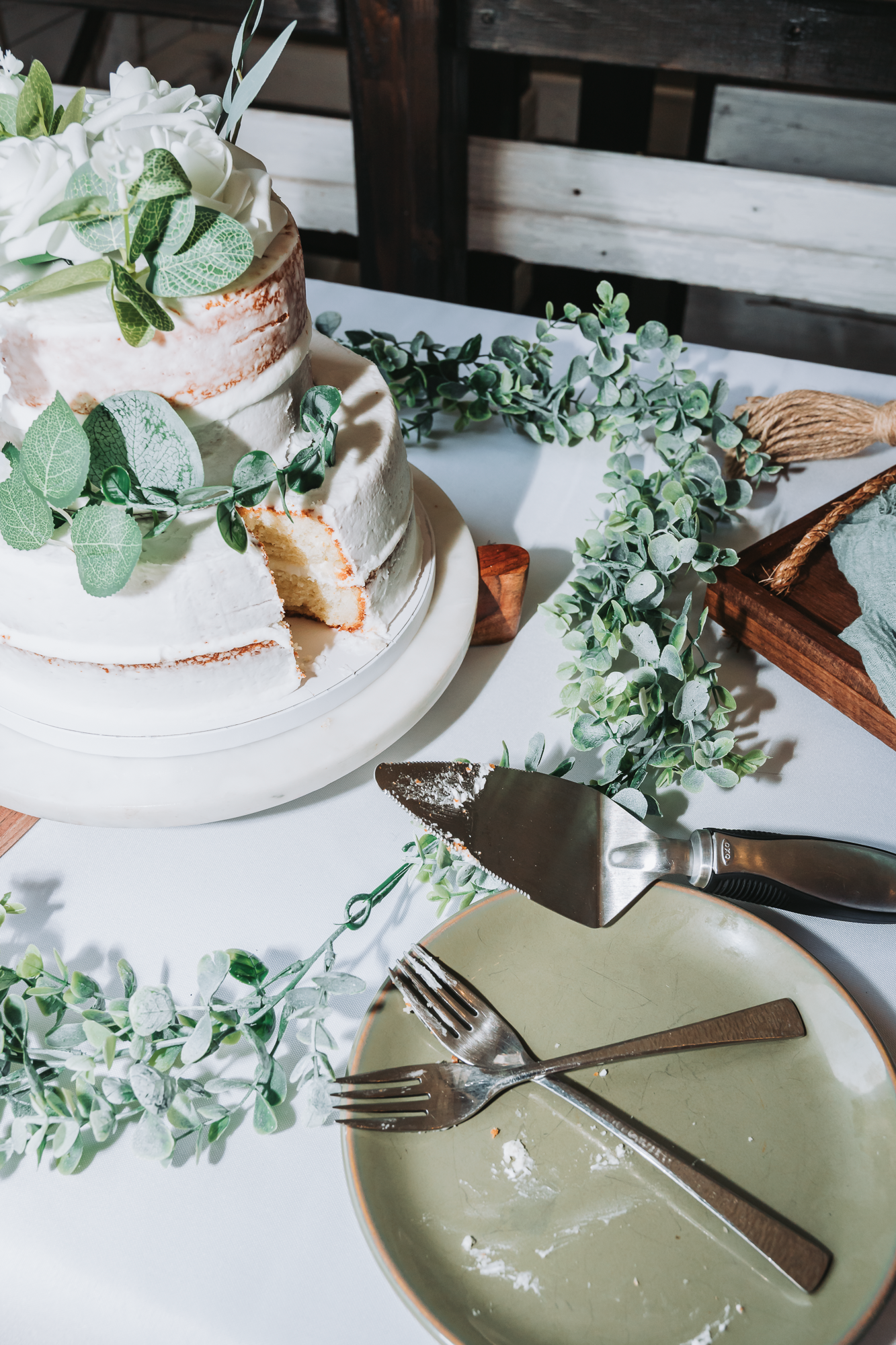 White wedding cake decorated with eucalyptus leaves and flowers on a rustic table setting.