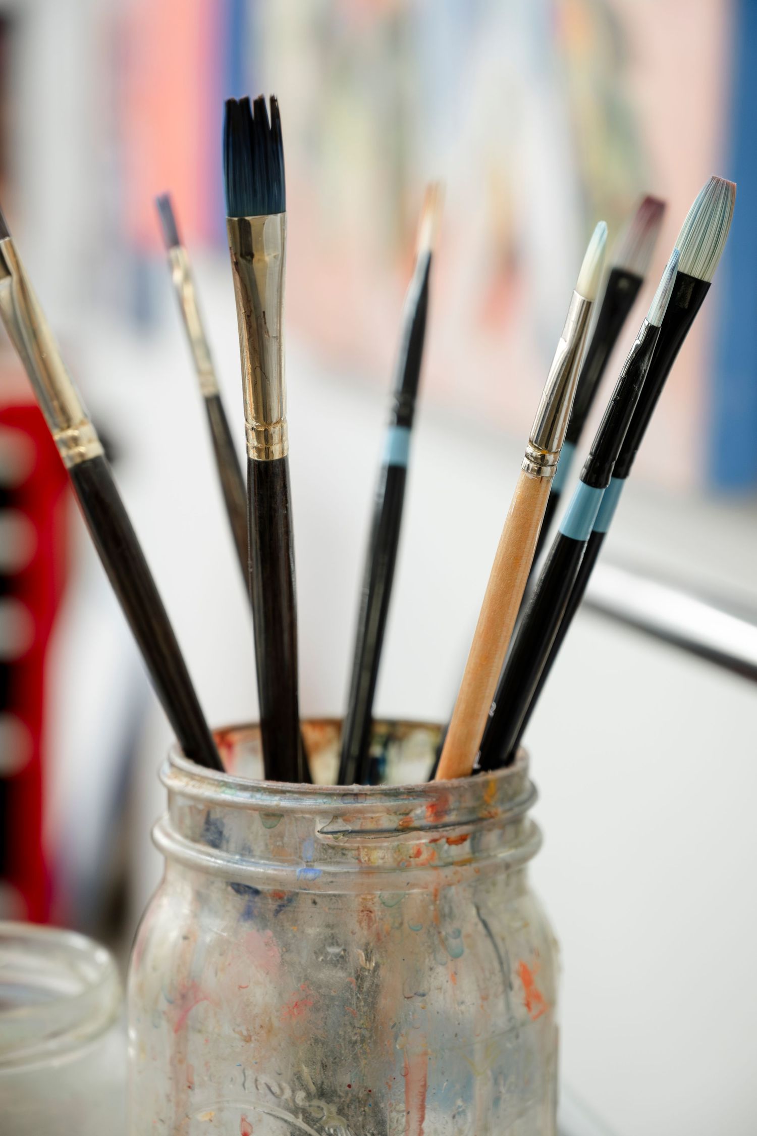 Paint brushes standing upright in a paint-stained glass jar against a blurred background.