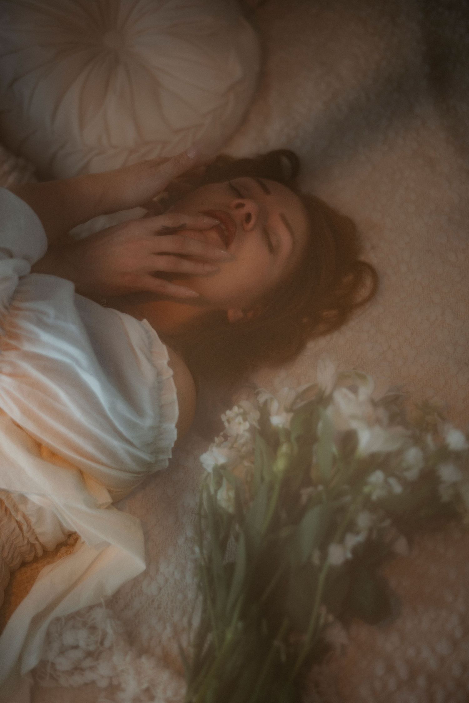 A woman in a white dress lies on a soft carpet, hand over her mouth, surrounded by white flowers and pillows.