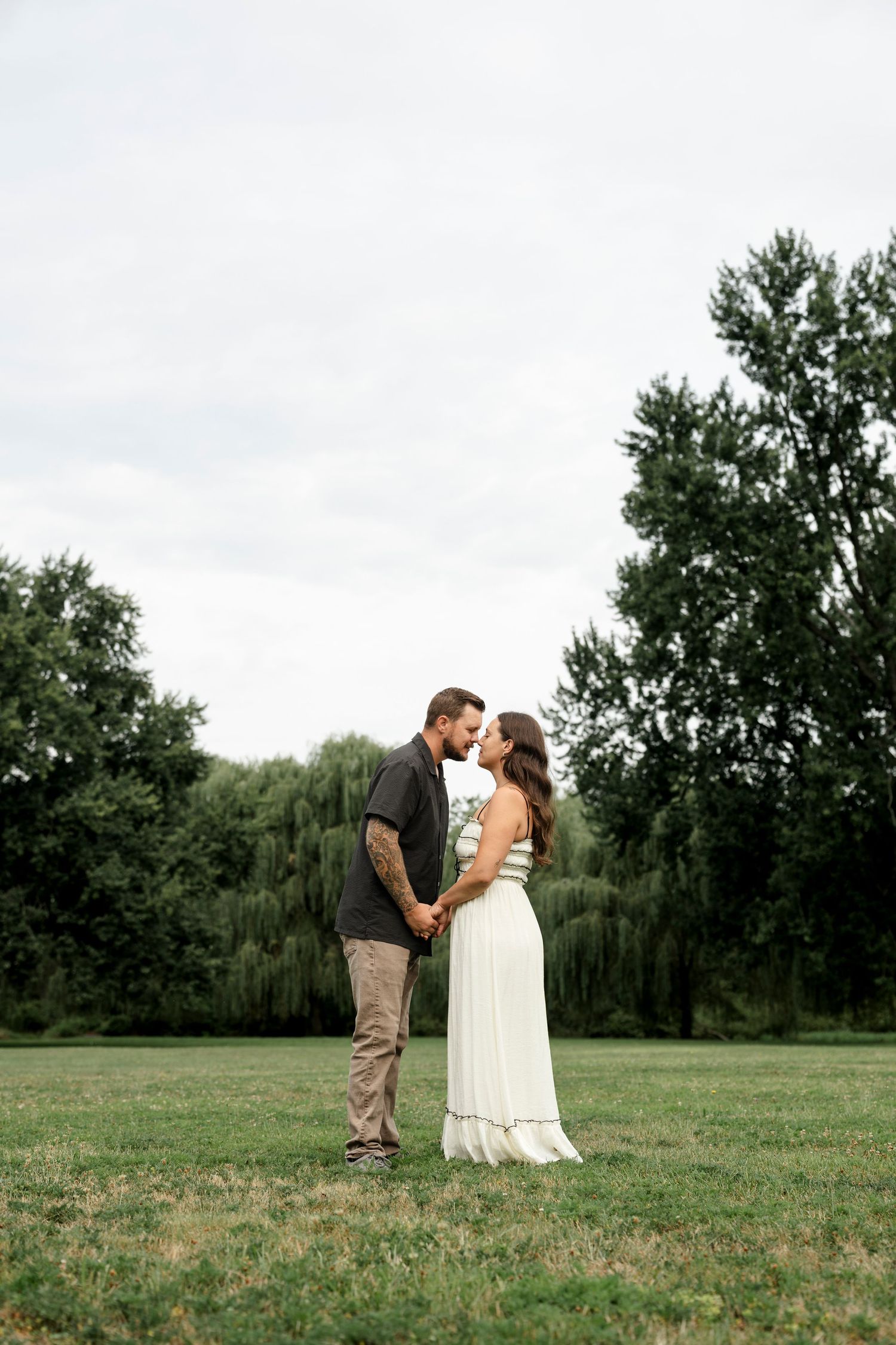 Couple sharing a romantic moment holding hands in a lush green park, woman in white maxi dress, man in dark shirt.