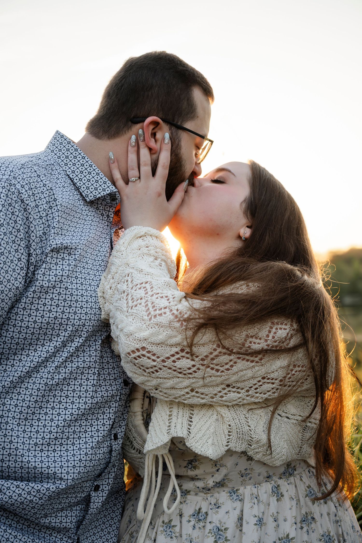A couple shares a romantic kiss outdoors at golden hour, the woman in a lace sweater gently holding his face.