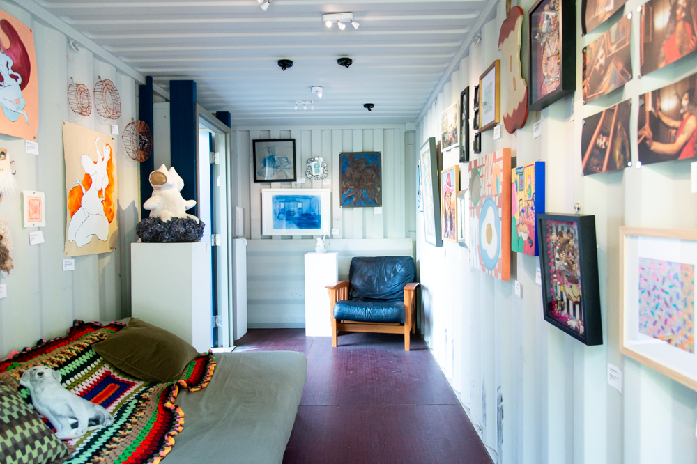 Art gallery hallway with colorful paintings displayed on white walls and a blue armchair.