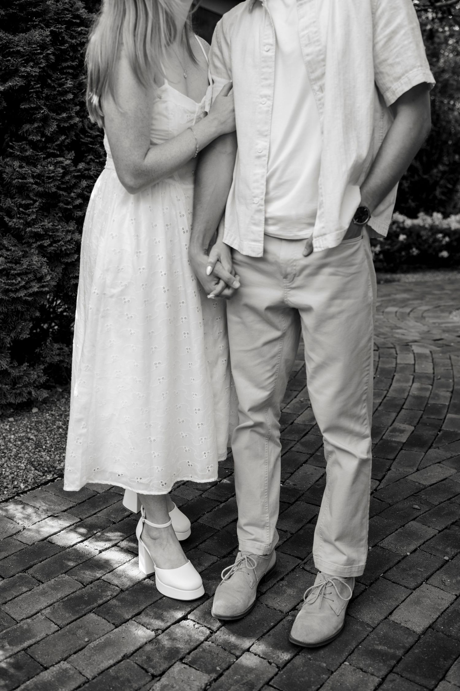 Black and white photo of a couple holding hands, woman in white dress, man in light shirt and pants, standing on brick path.