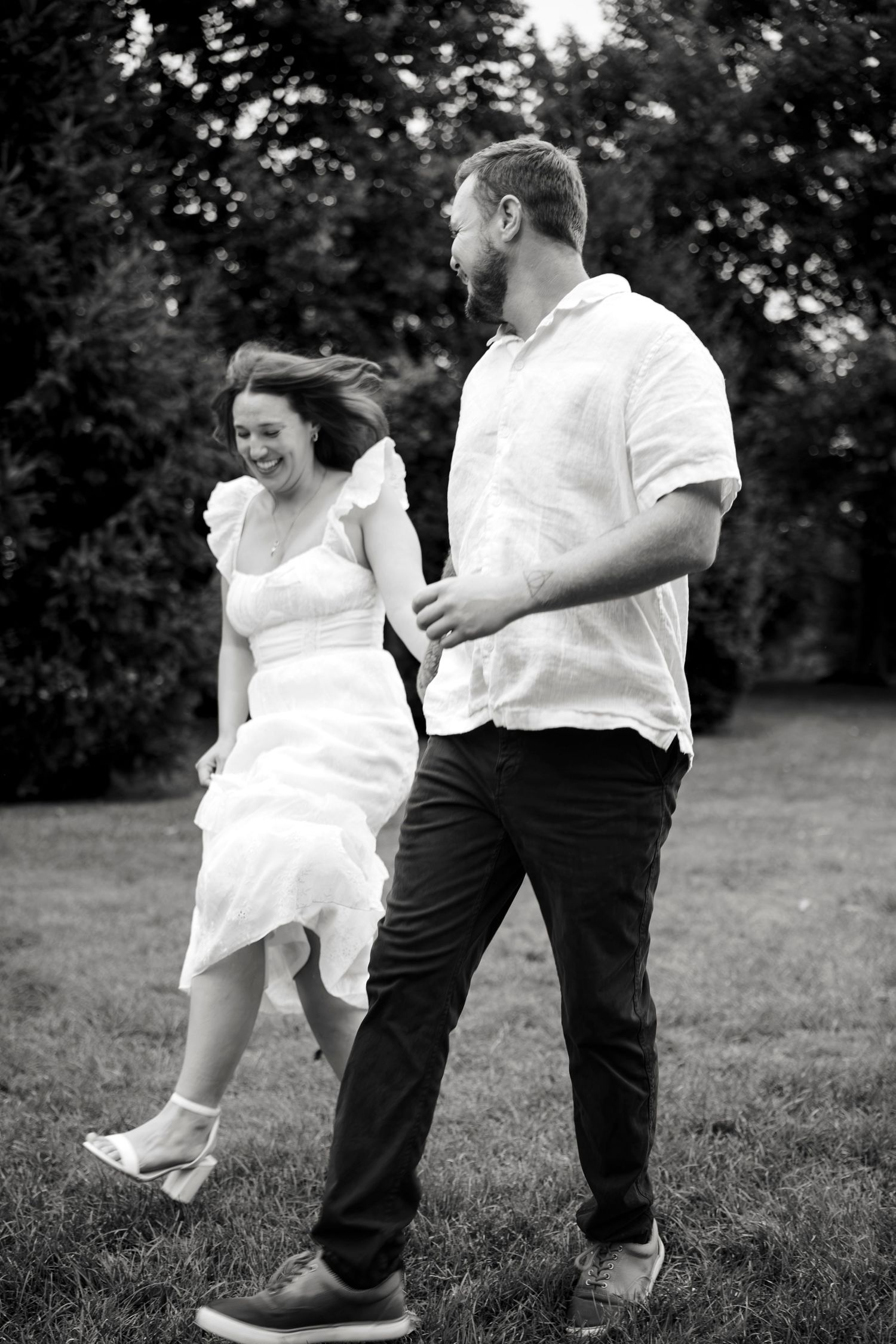 Black and white photo of a laughing couple walking hand in hand outdoors, woman in white dress, man in white shirt.