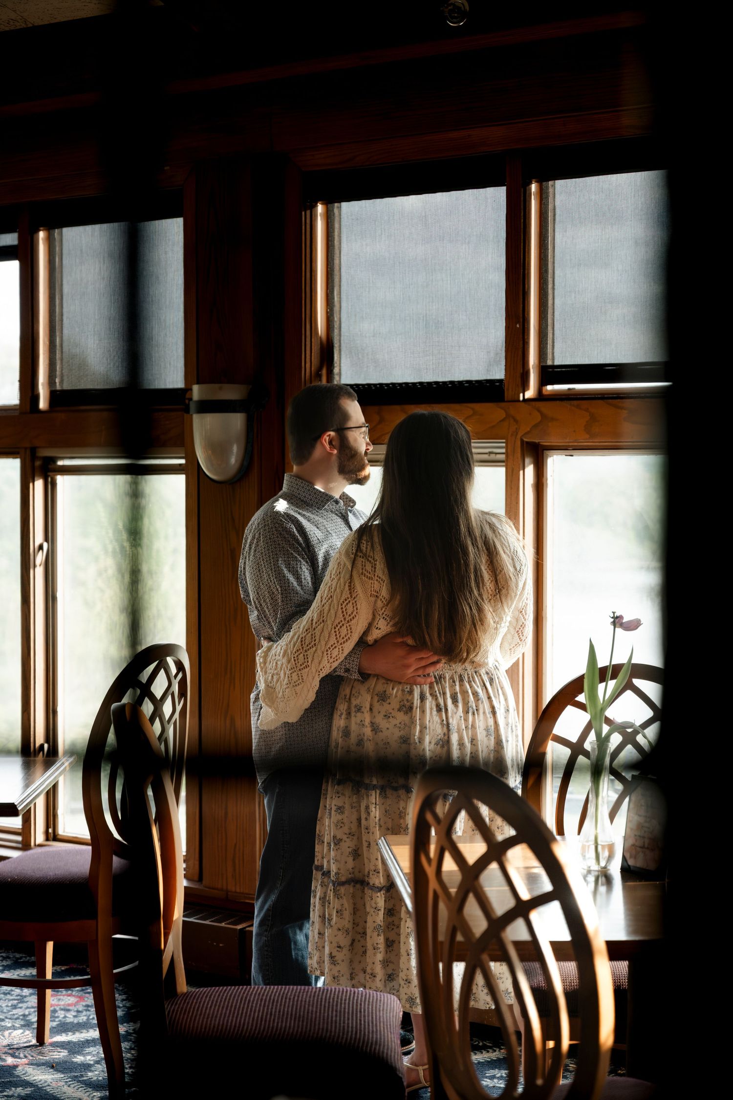 Couple embracing near sunlit windows in a warmly lit restaurant with wooden chairs and elegant decor.