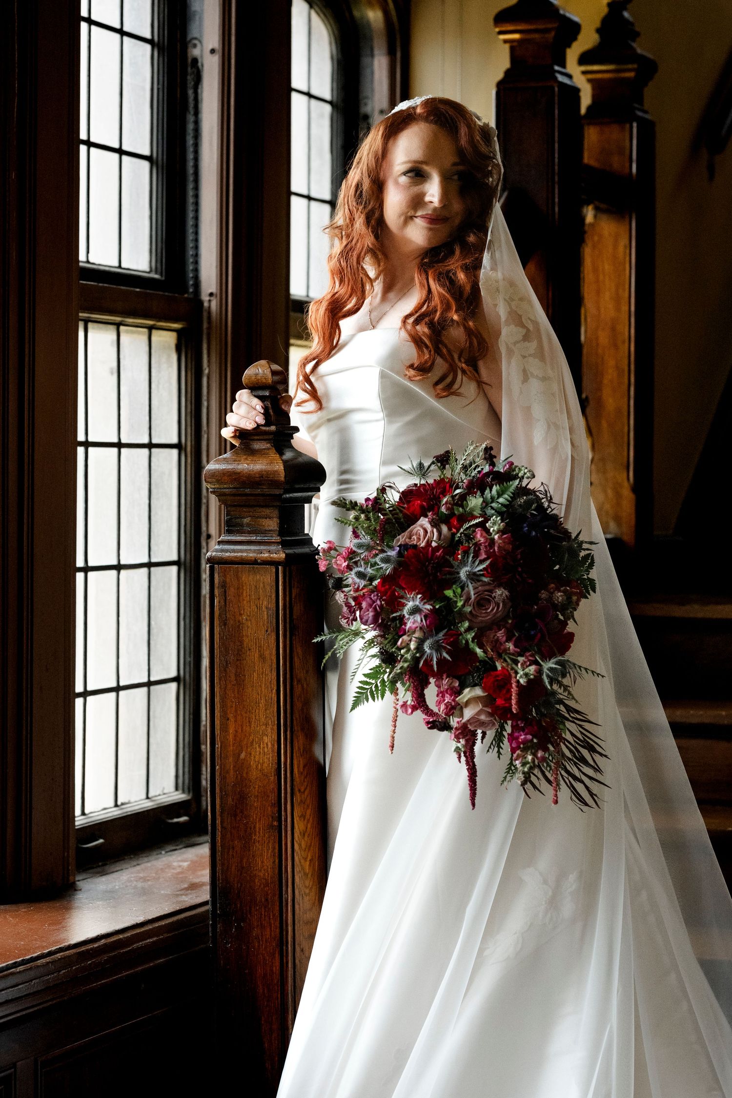 A bride in a white ballgown holds a deep burgundy and blush floral bouquet while standing by a window in a rustic venue.