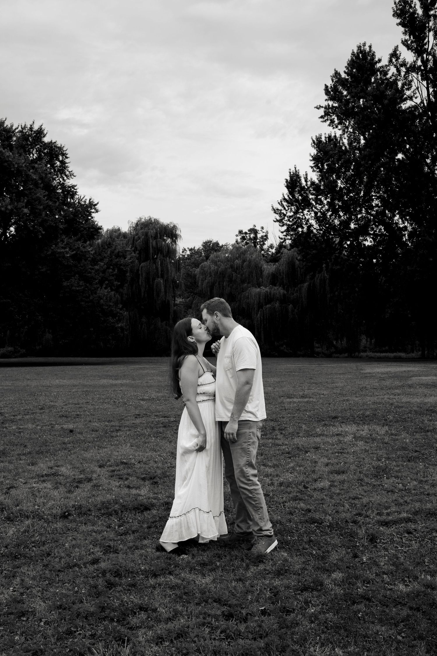 Black and white photo of a couple sharing a romantic kiss in a lush green park surrounded by tall trees.