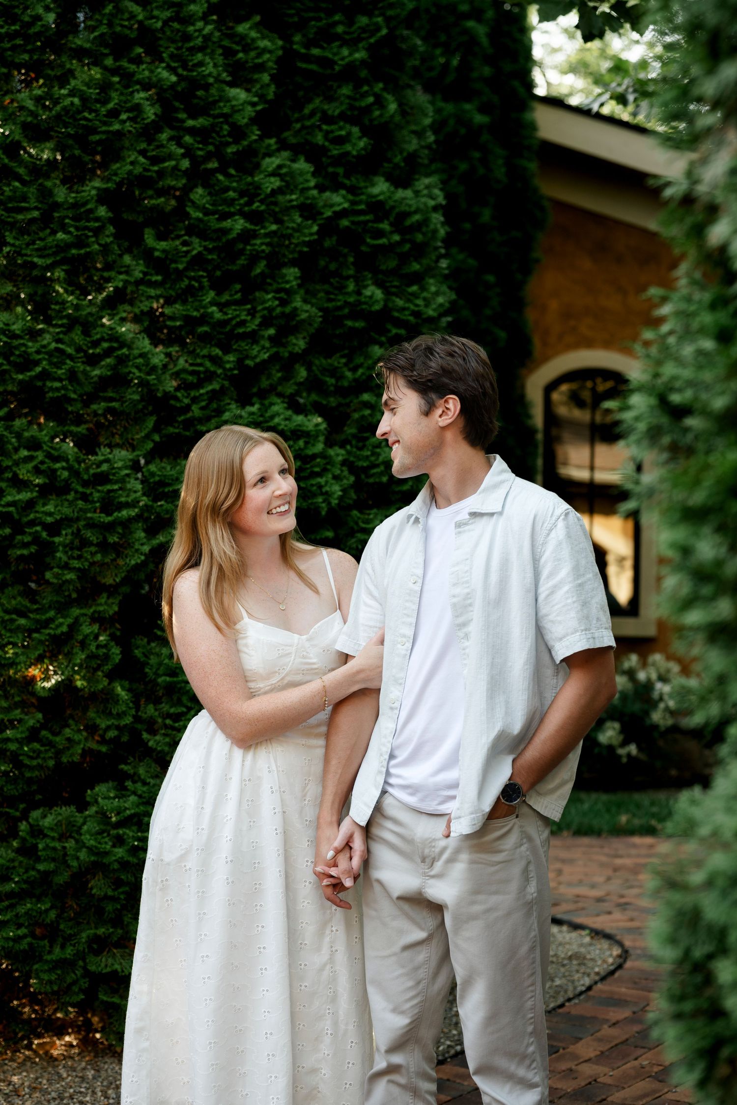 A smiling couple in white outfits hold hands outdoors, surrounded by lush green trees near a brick building.