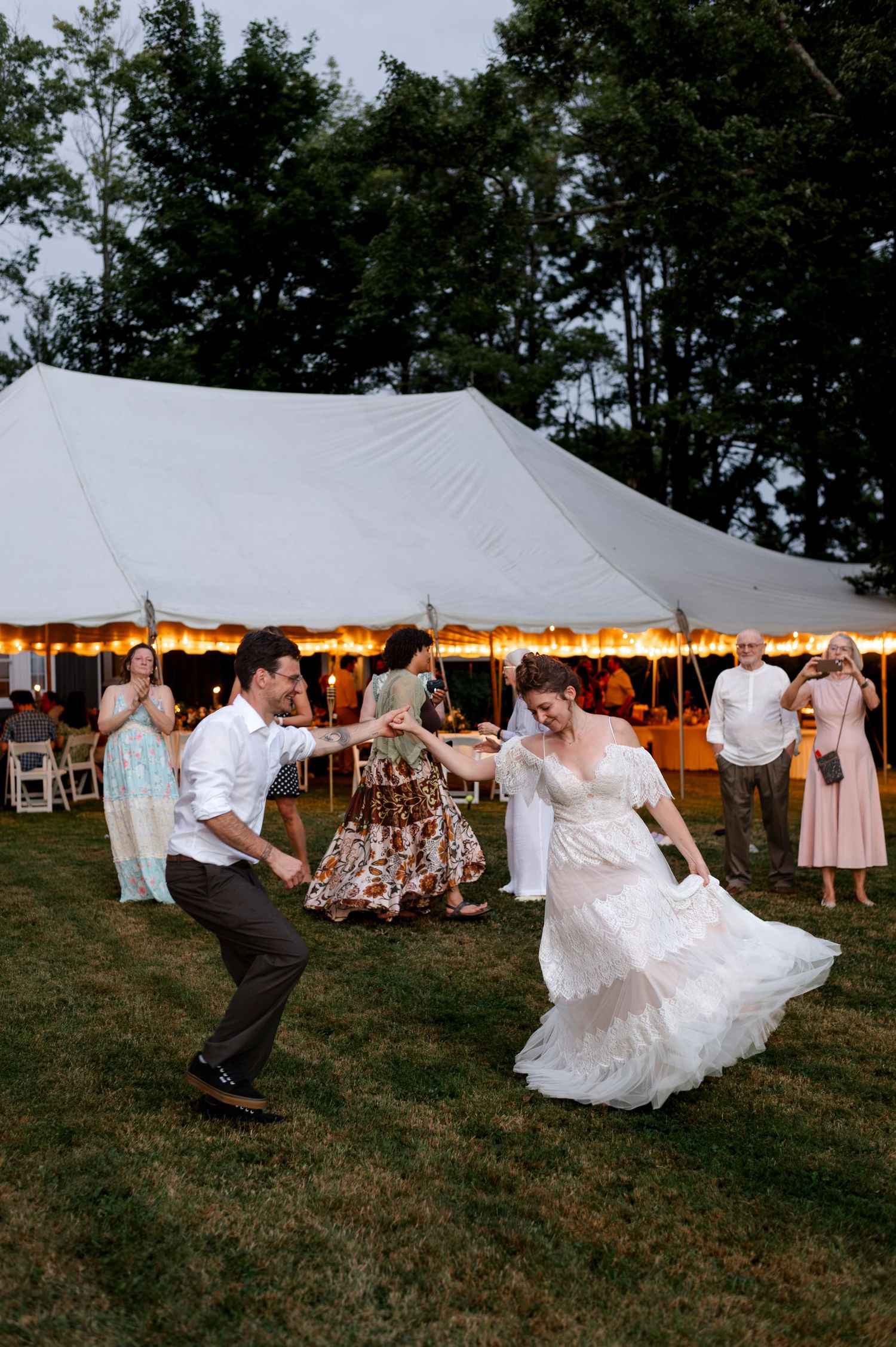 Bride and groom dancing outdoors at a tented wedding reception on a lush green lawn at dusk.