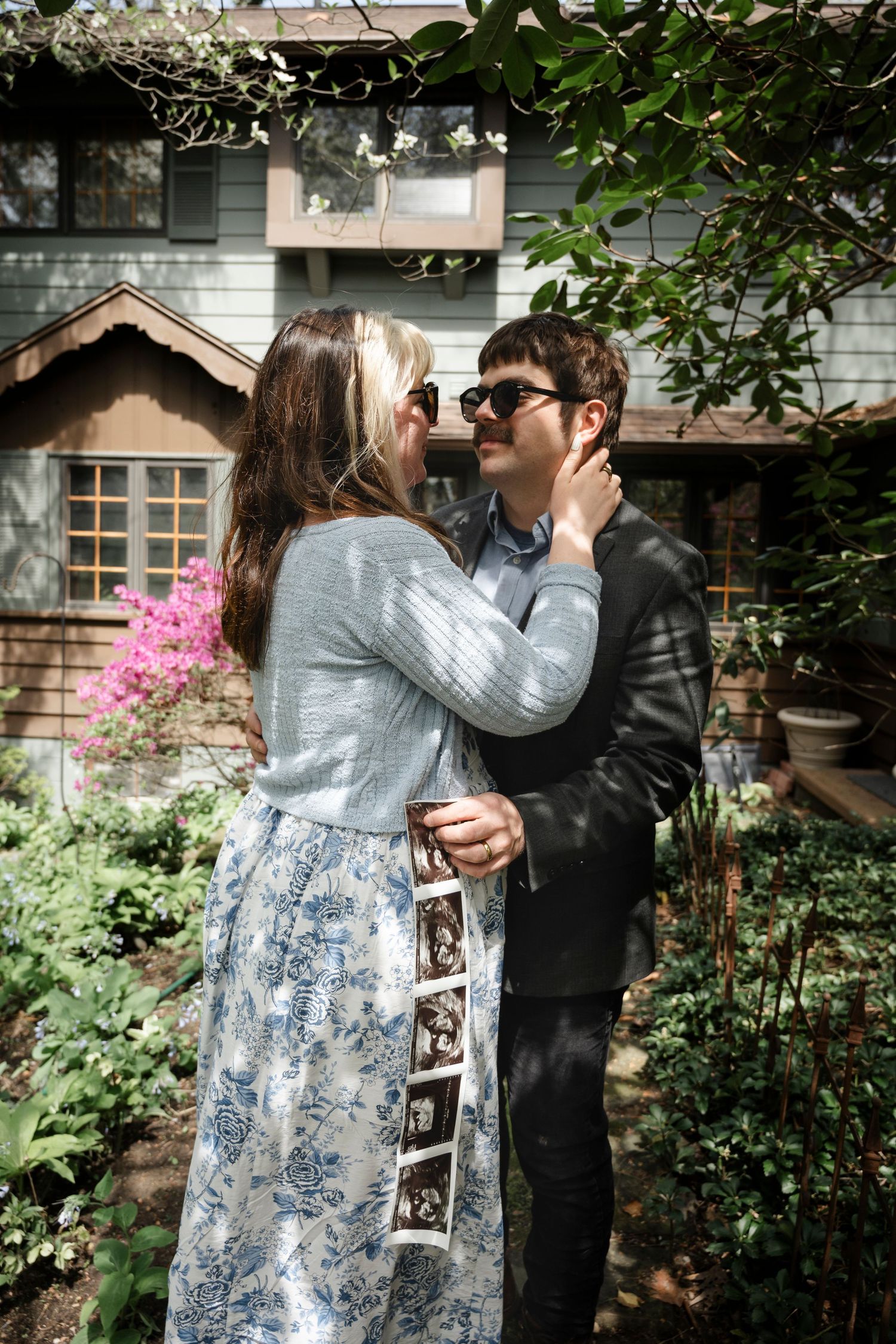 Couple embracing in garden holding ultrasound photos, announcing pregnancy outside a charming house.