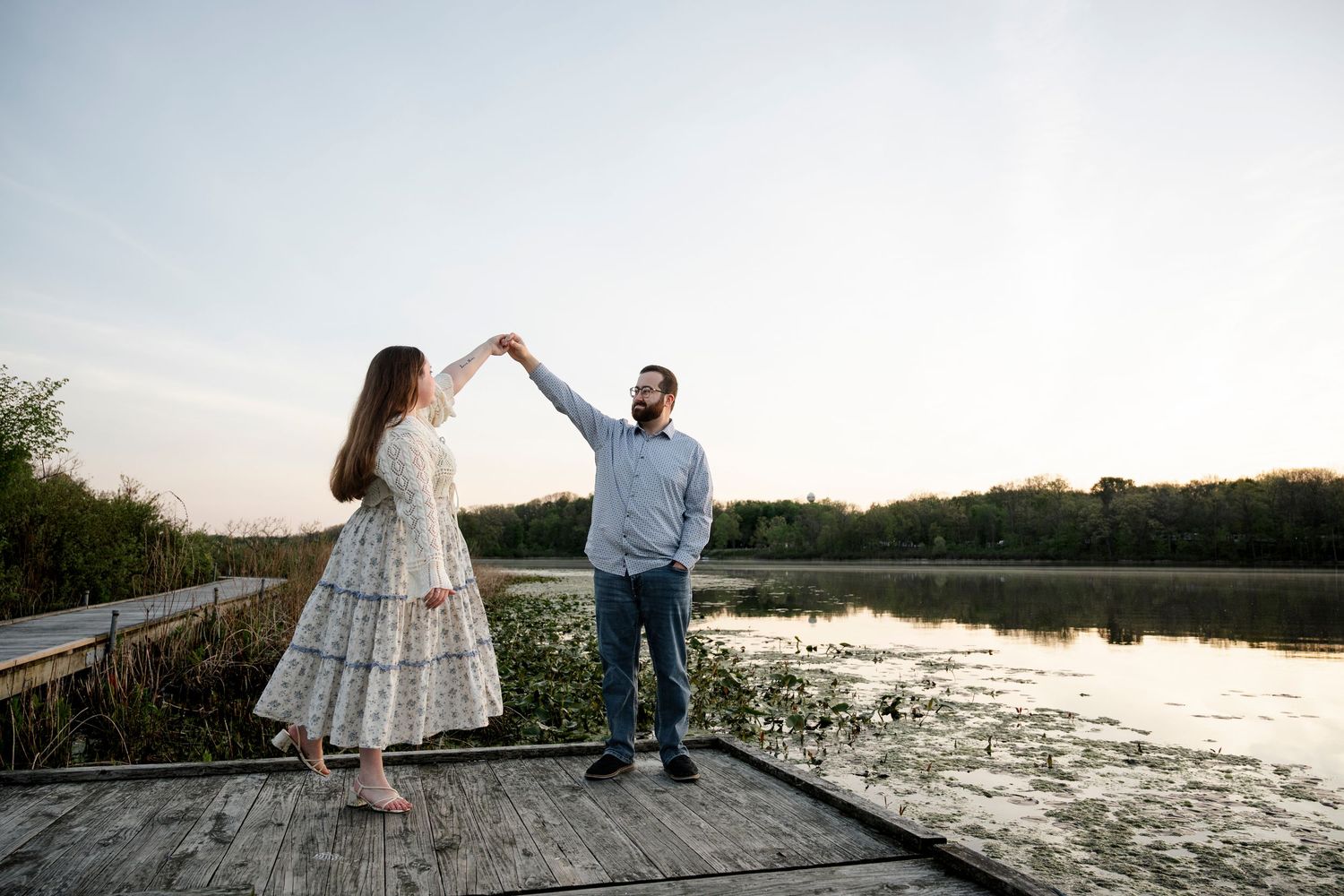 Couple dancing on a wooden dock by a serene lake at sunset, surrounded by lush green trees.