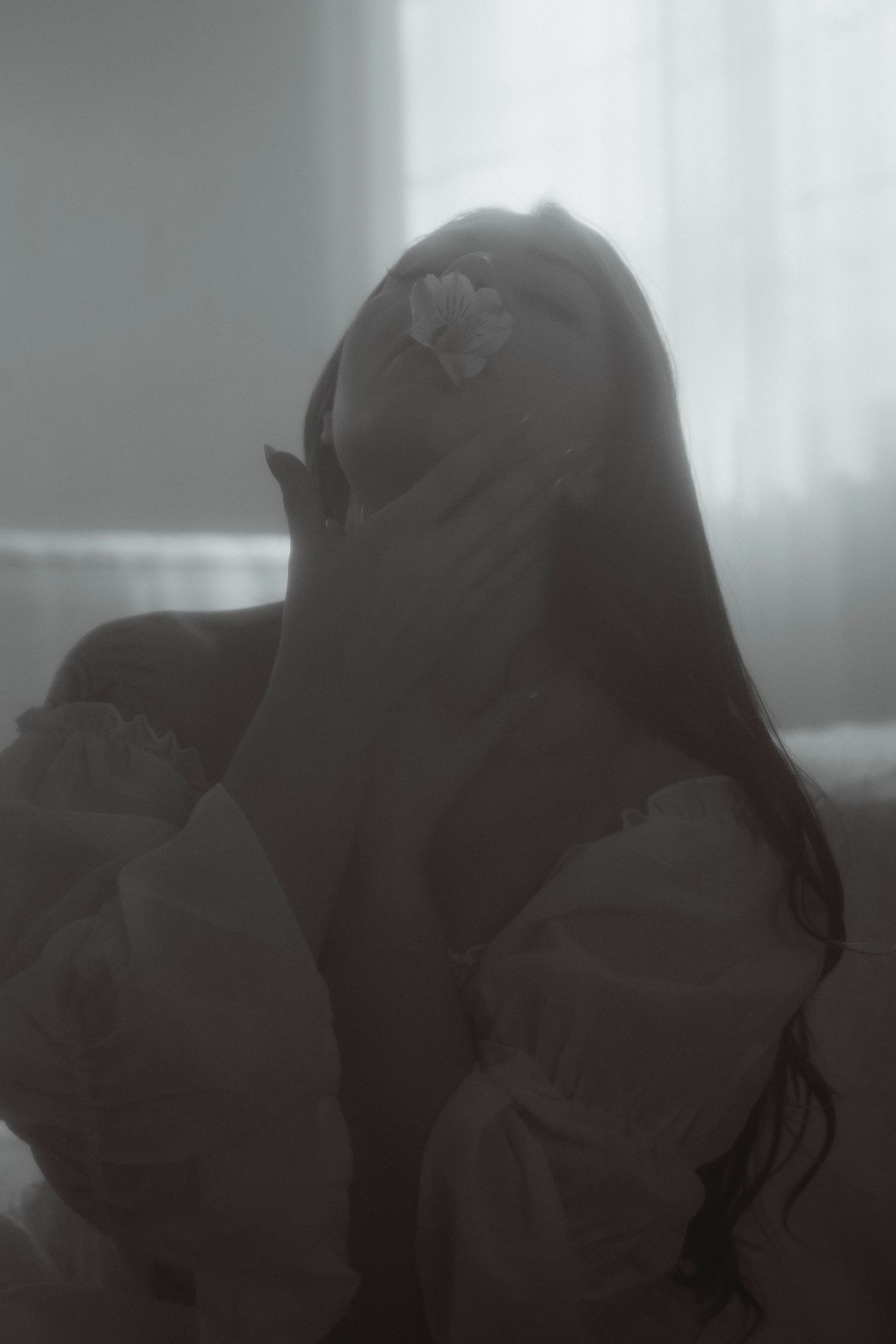 Moody monochrome portrait of woman with flower in hair covering face on bed.