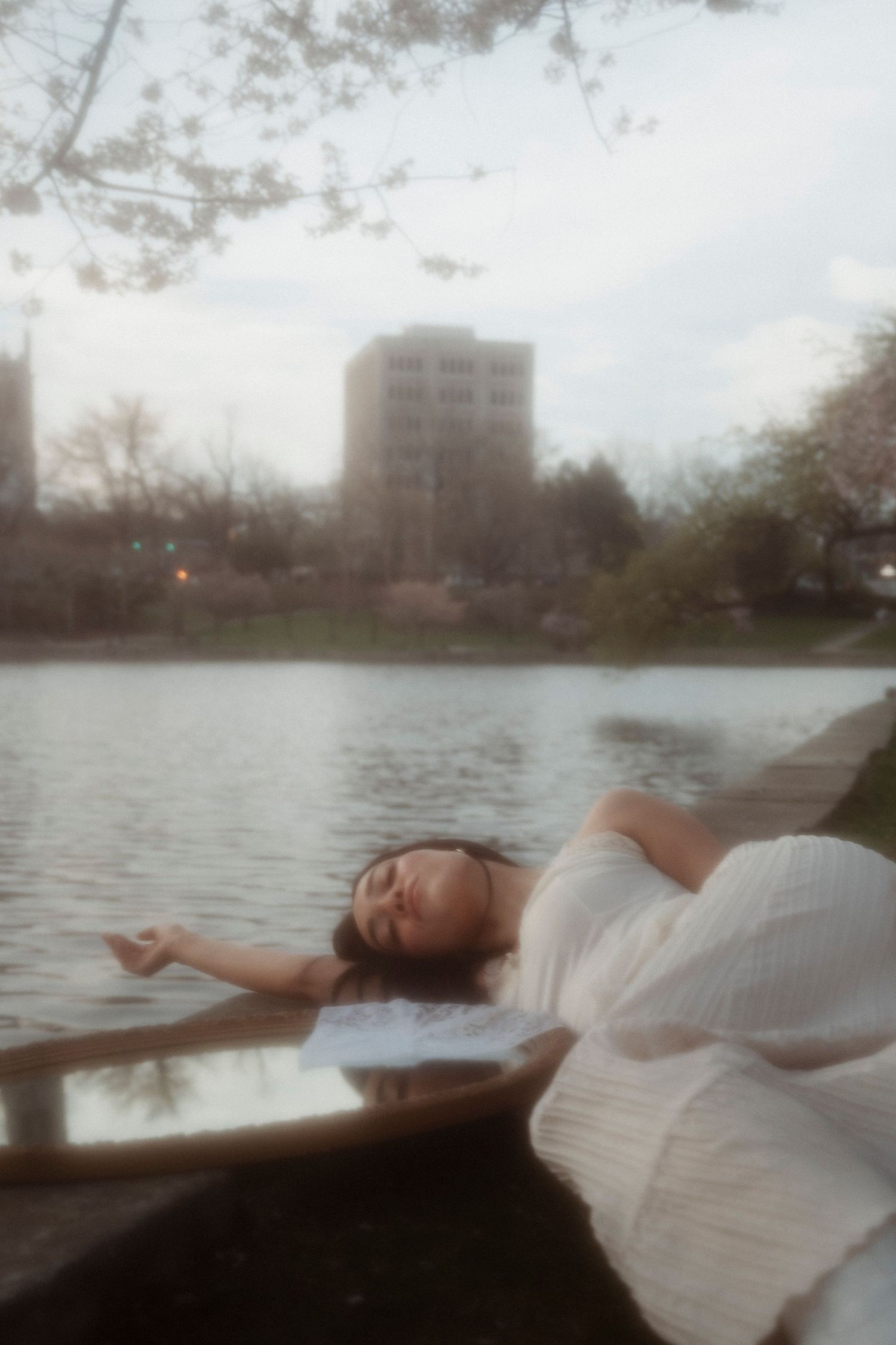 Woman in white dress reclines peacefully in a boat on a serene urban lake surrounded by cherry blossoms.