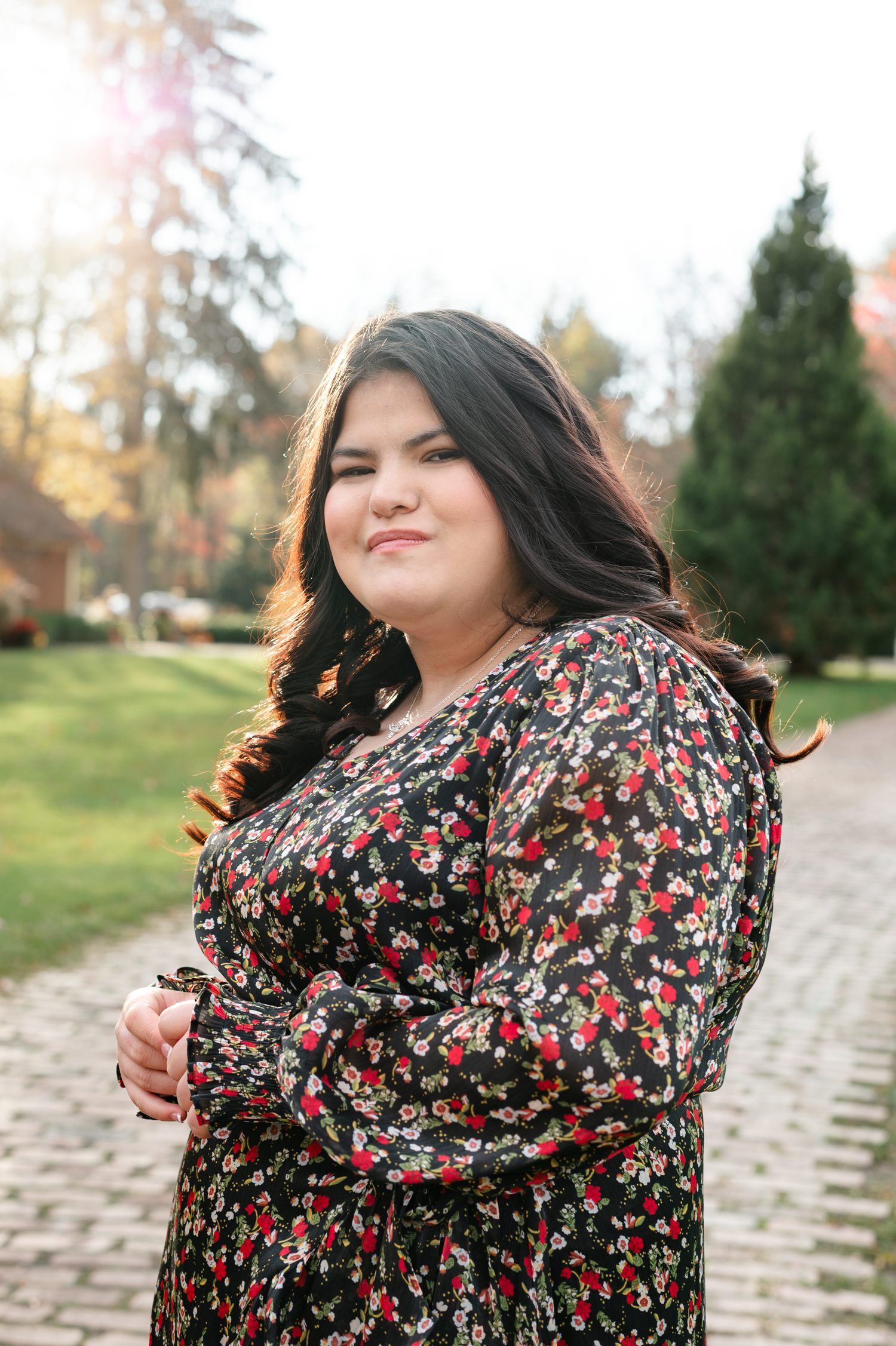 A person in a floral print dress poses outdoors on a stone path with autumn trees in the background.