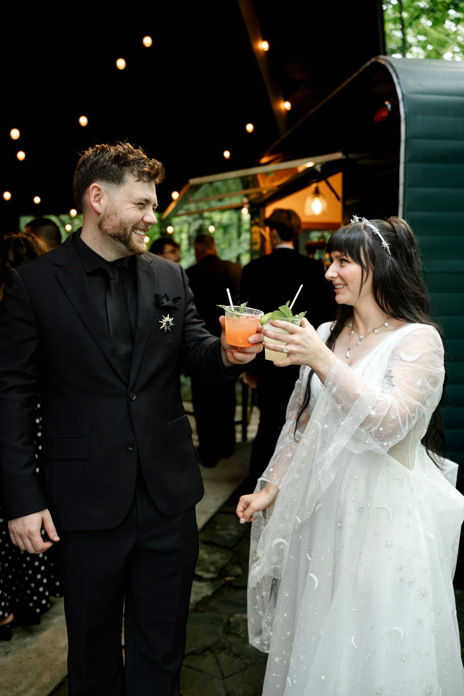 Bride and groom toasting with cocktails at outdoor wedding reception, string lights glowing behind them.