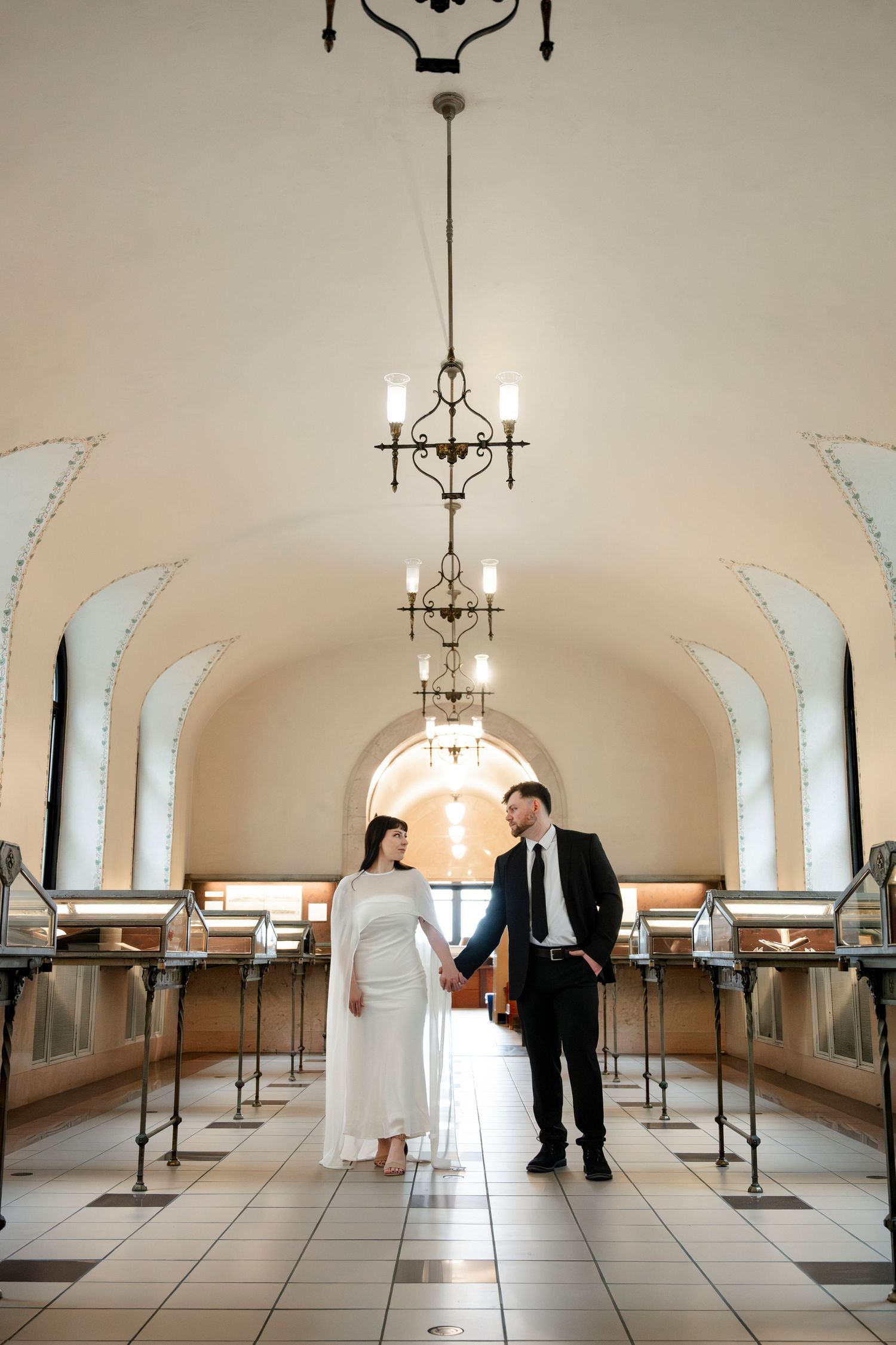 Elegant couple in formal attire holding hands in a grand hall with arched ceilings and chandelier lighting.