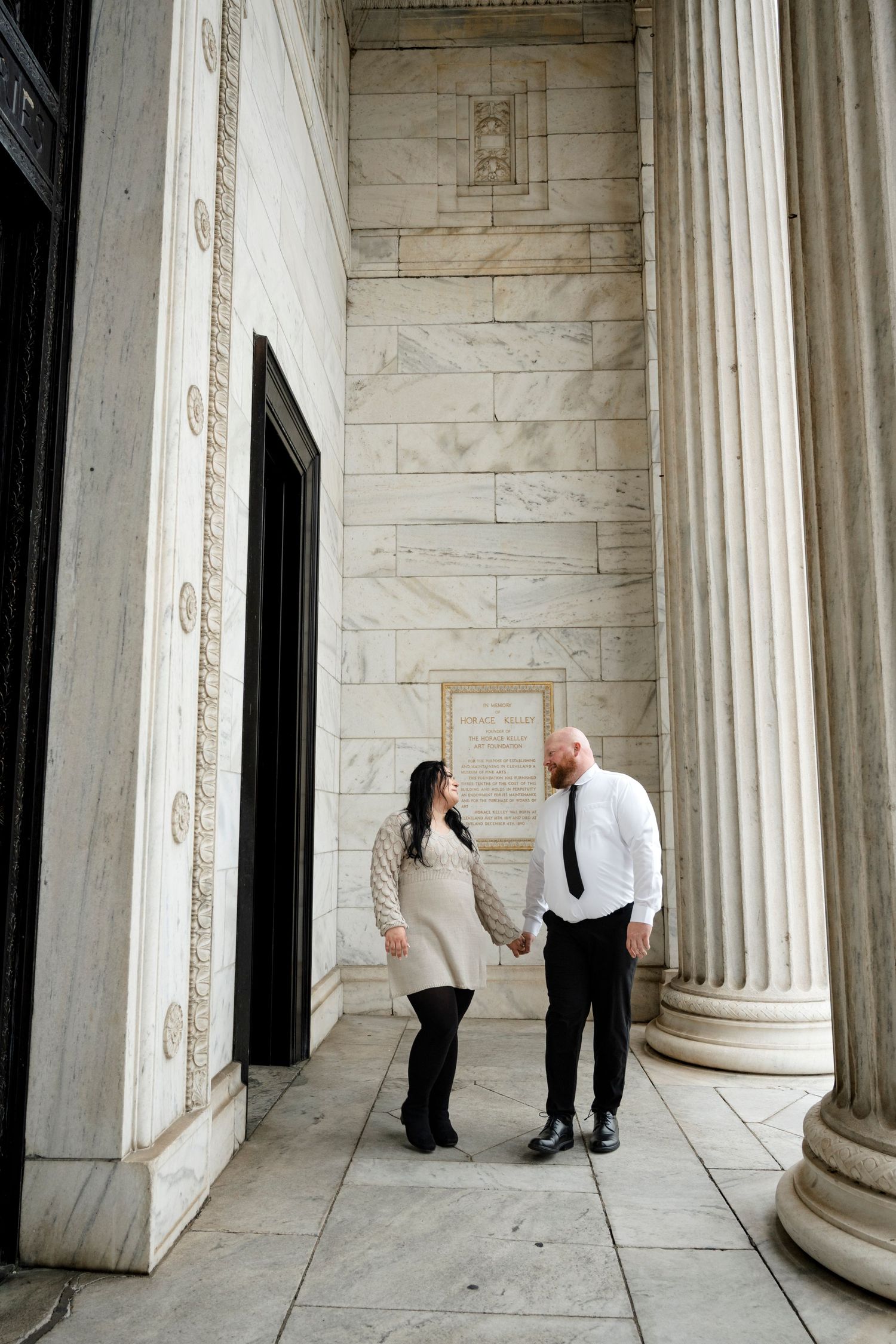 Couple holding hands and smiling at each other beside marble columns of a grand building.