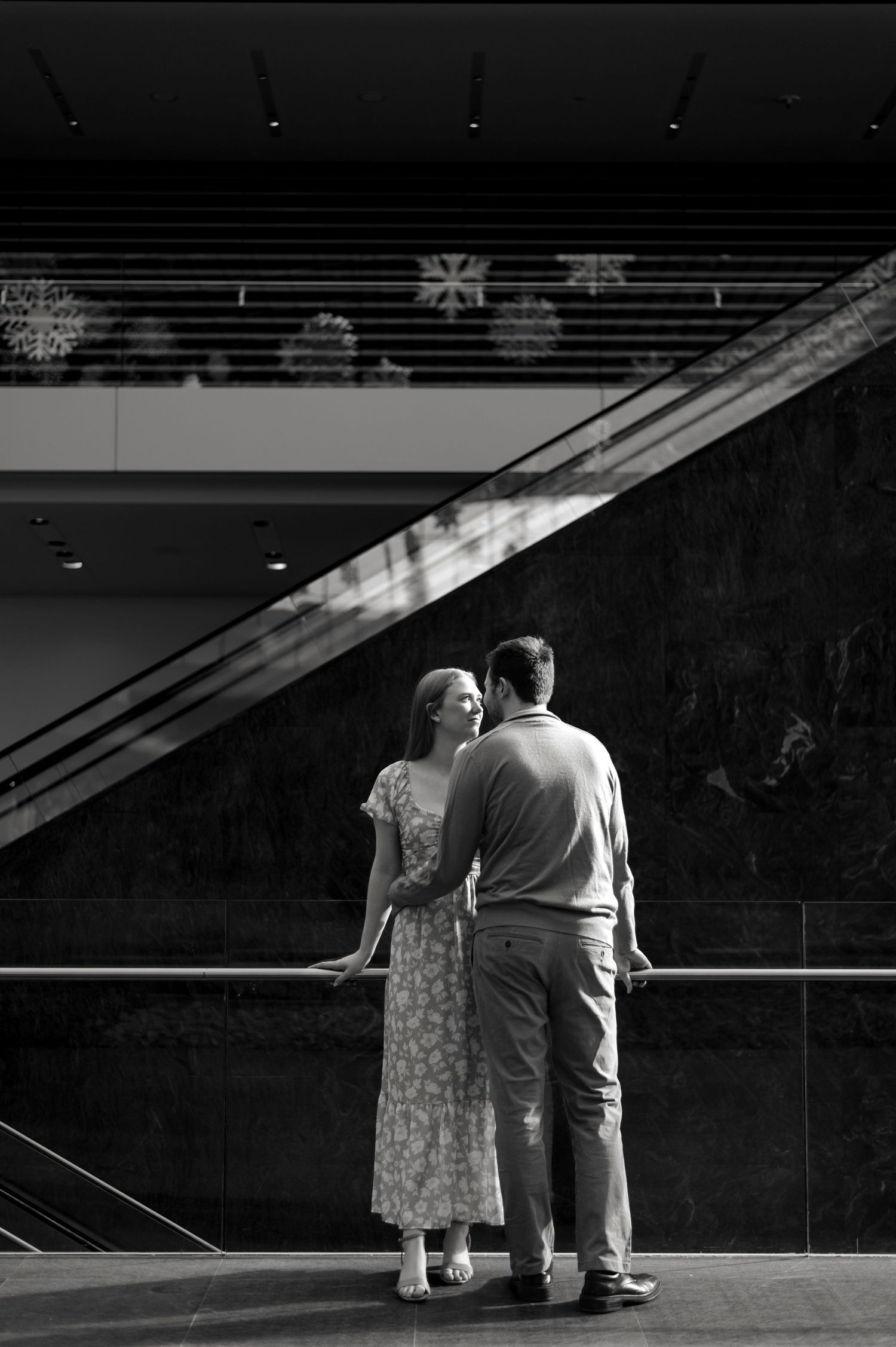 Black and white photo of a couple holding hands, standing together near a railing inside an elegant building.