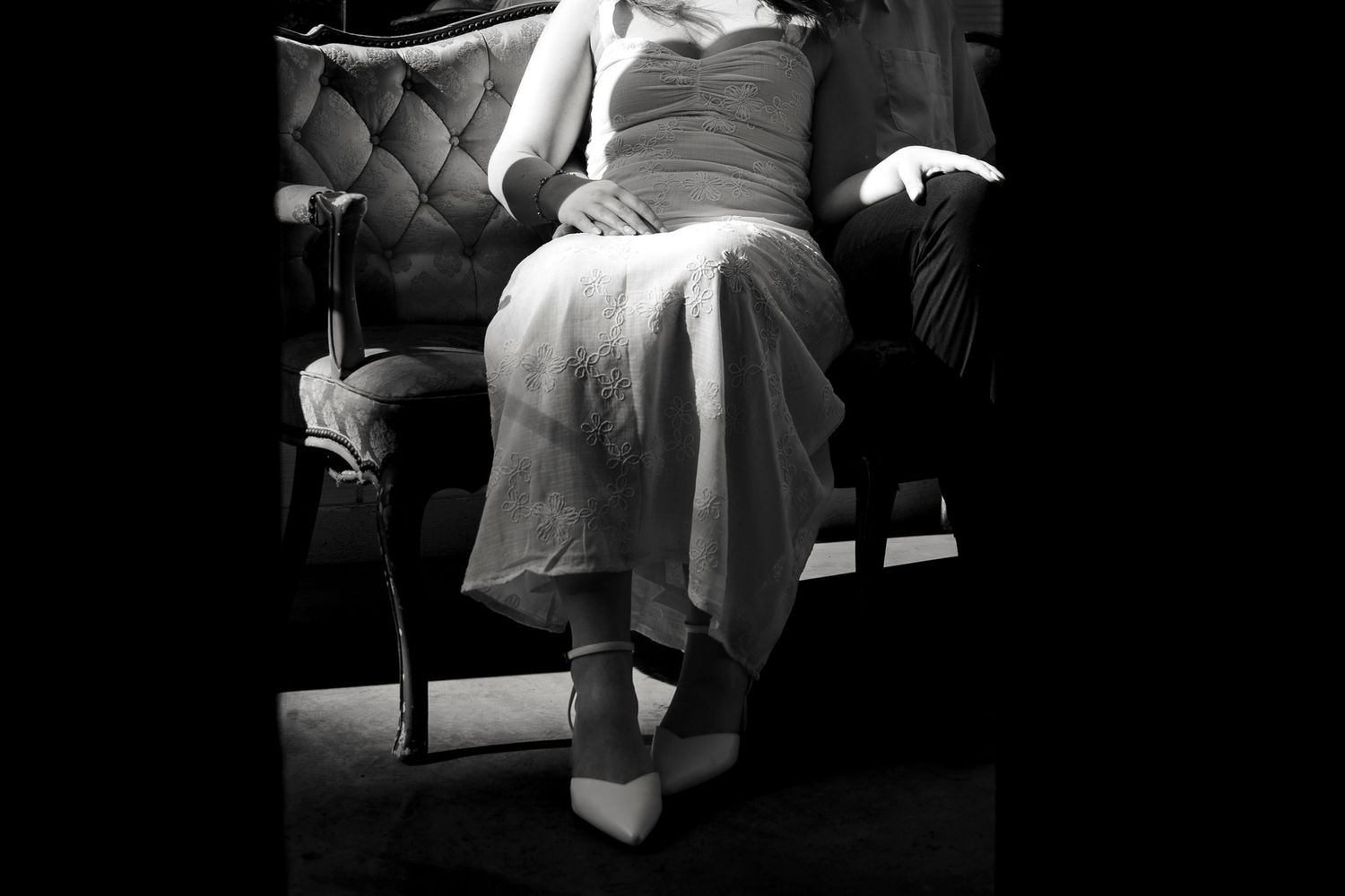 Black and white photo of a bride in a white gown and heels seated elegantly in a vintage chair.