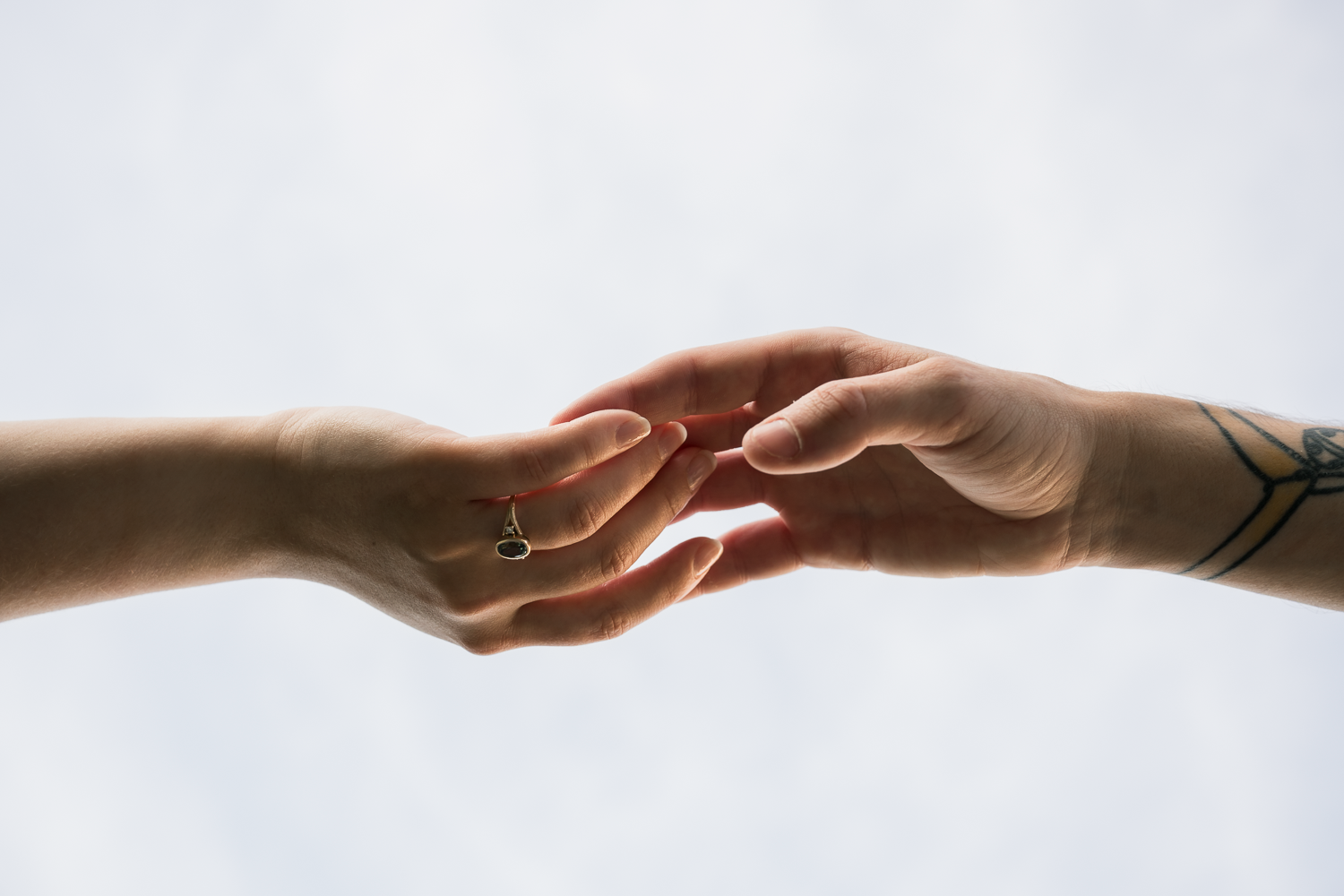 Two hands gently touching fingertips, one wearing a ring and the other with a tattoo, against a white background.