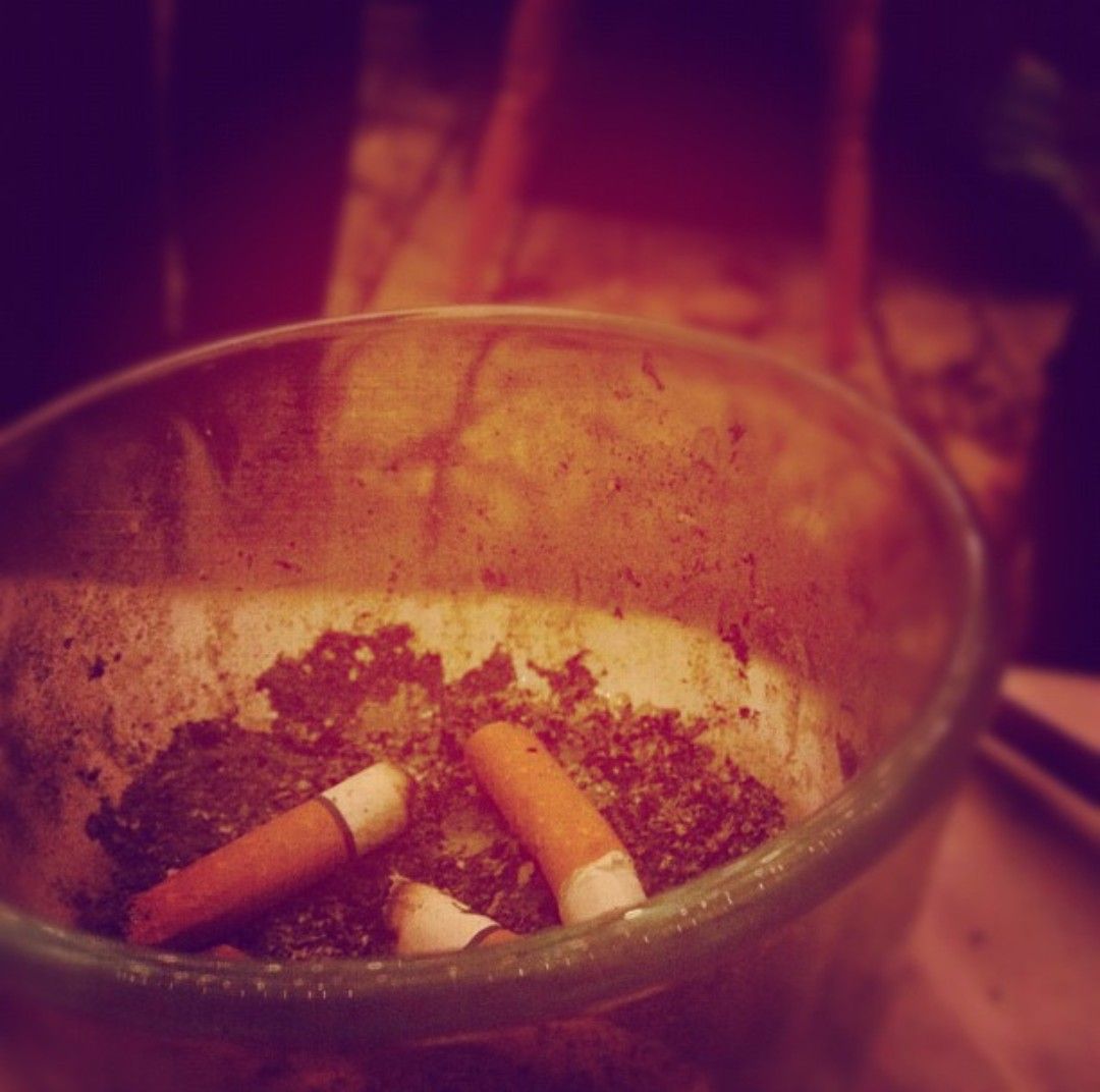 Cigarette butts discarded in a glass ashtray with ashes against a dark background.