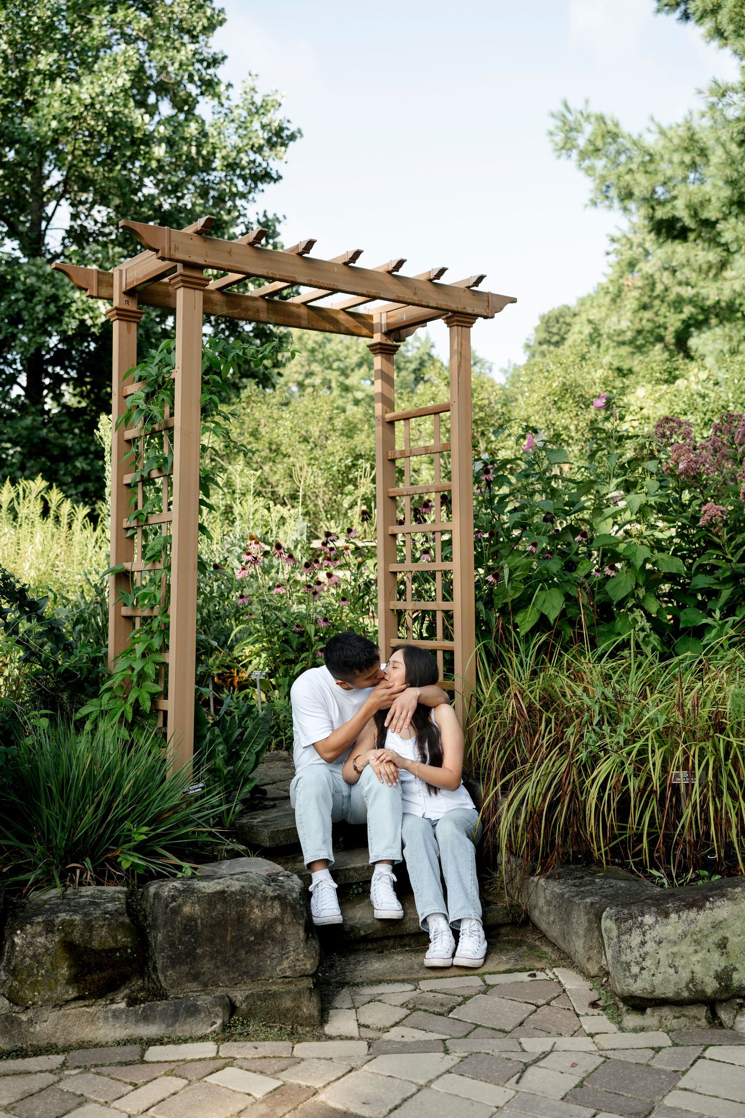 Couple sitting together on stone steps under a wooden pergola surrounded by lush green garden plants.