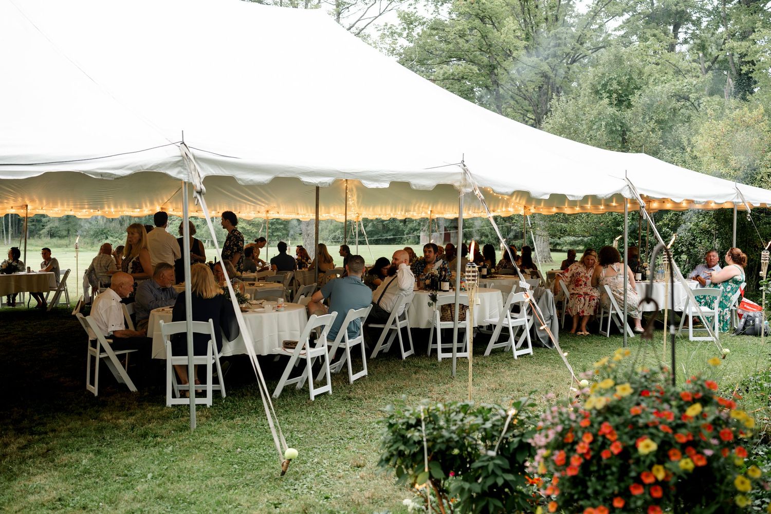 Guests dining under a white outdoor tent at a garden reception with white chairs and tables on green grass.