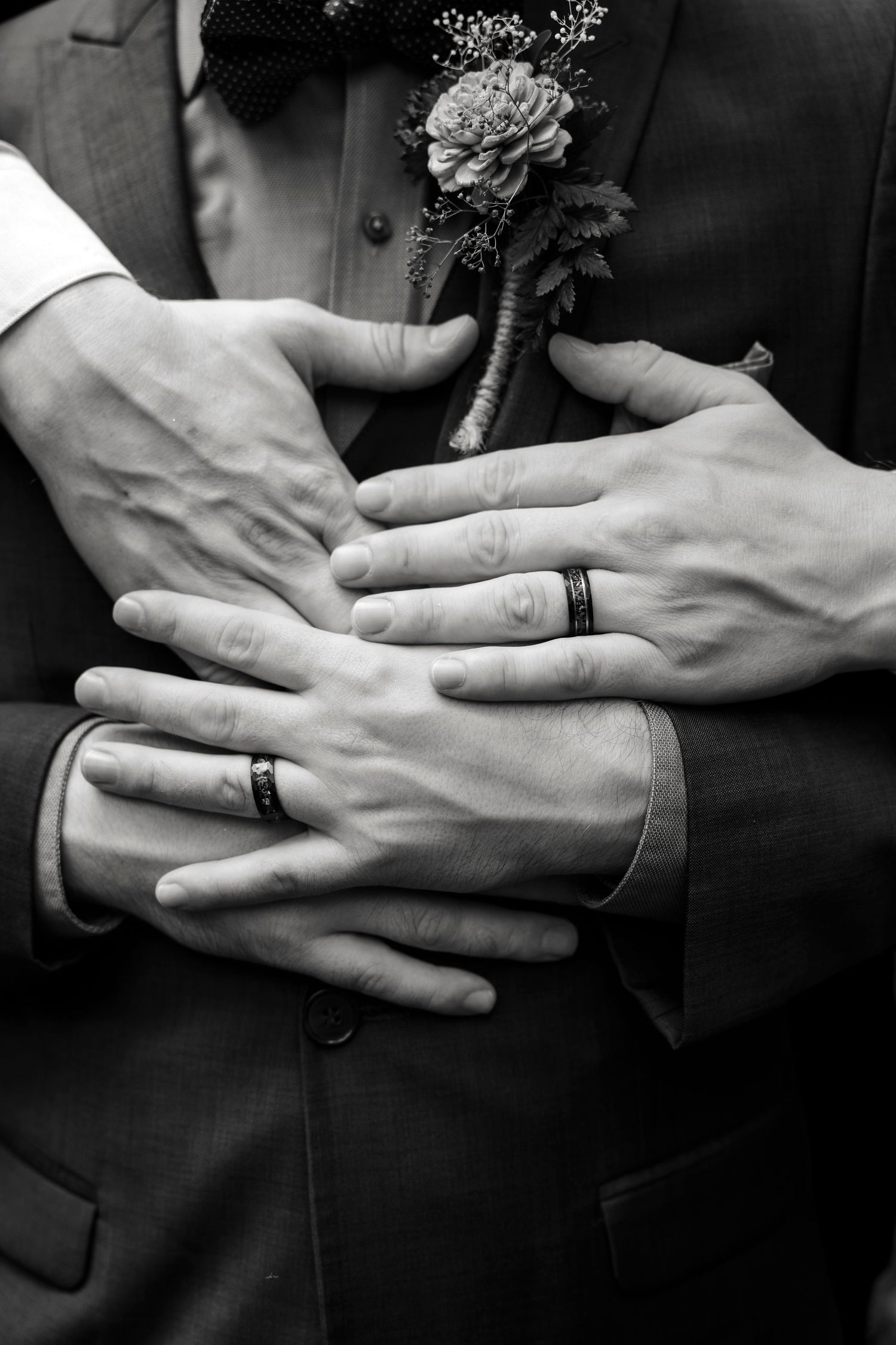 Black and white photo of two grooms embracing, showing wedding rings on their hands with a boutonniere visible.