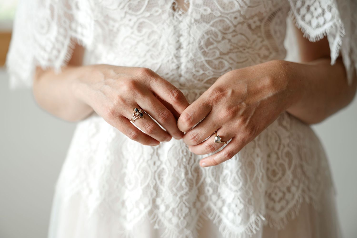 Close-up of a bride in white lace wedding dress clasping hands, wearing elegant gold rings.