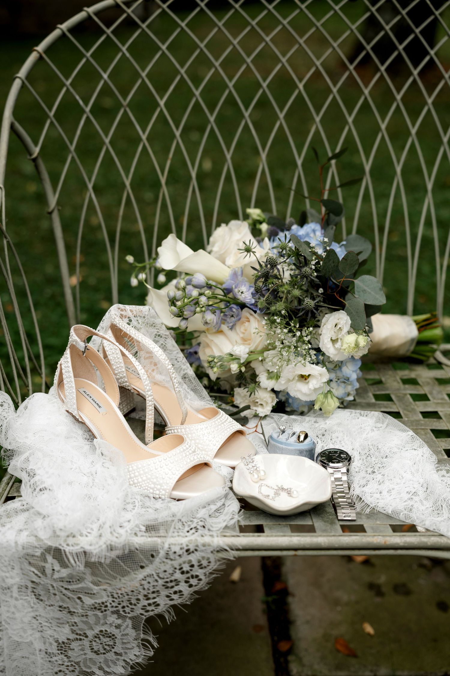 White bridal heels, lace veil, floral bouquet with blue and white flowers, and jewelry displayed on a vintage metal chair.