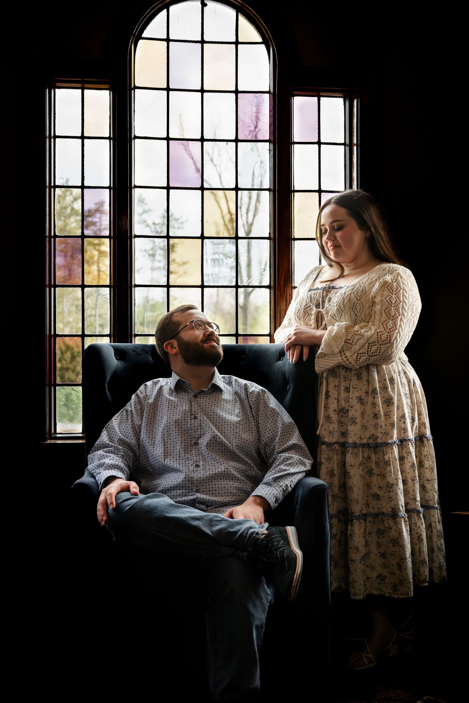 A couple poses indoors by arched windows, man seated in armchair, woman standing beside him in floral dress.