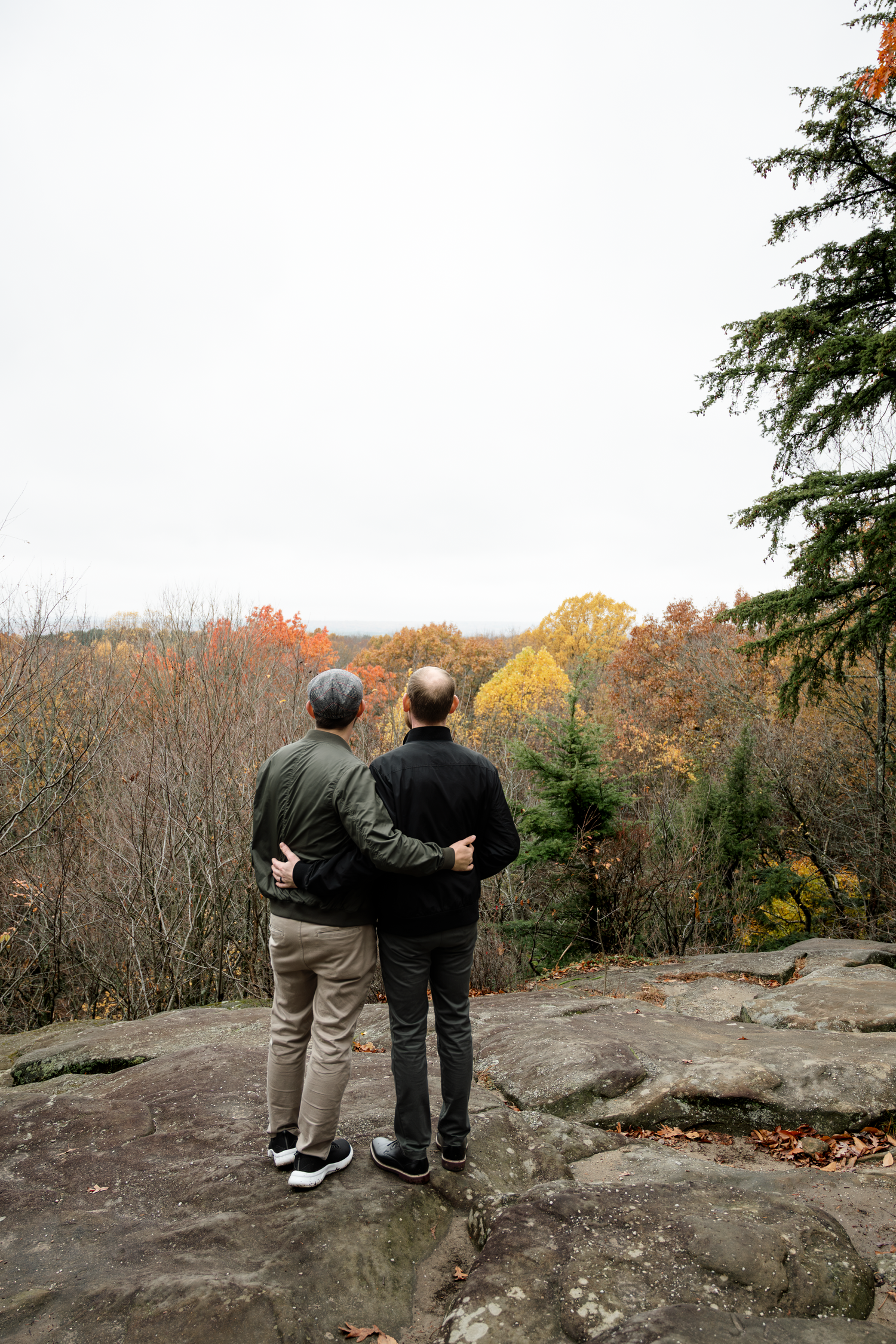 Two people embracing on a rocky overlook surrounded by colorful autumn foliage on an overcast fall day.