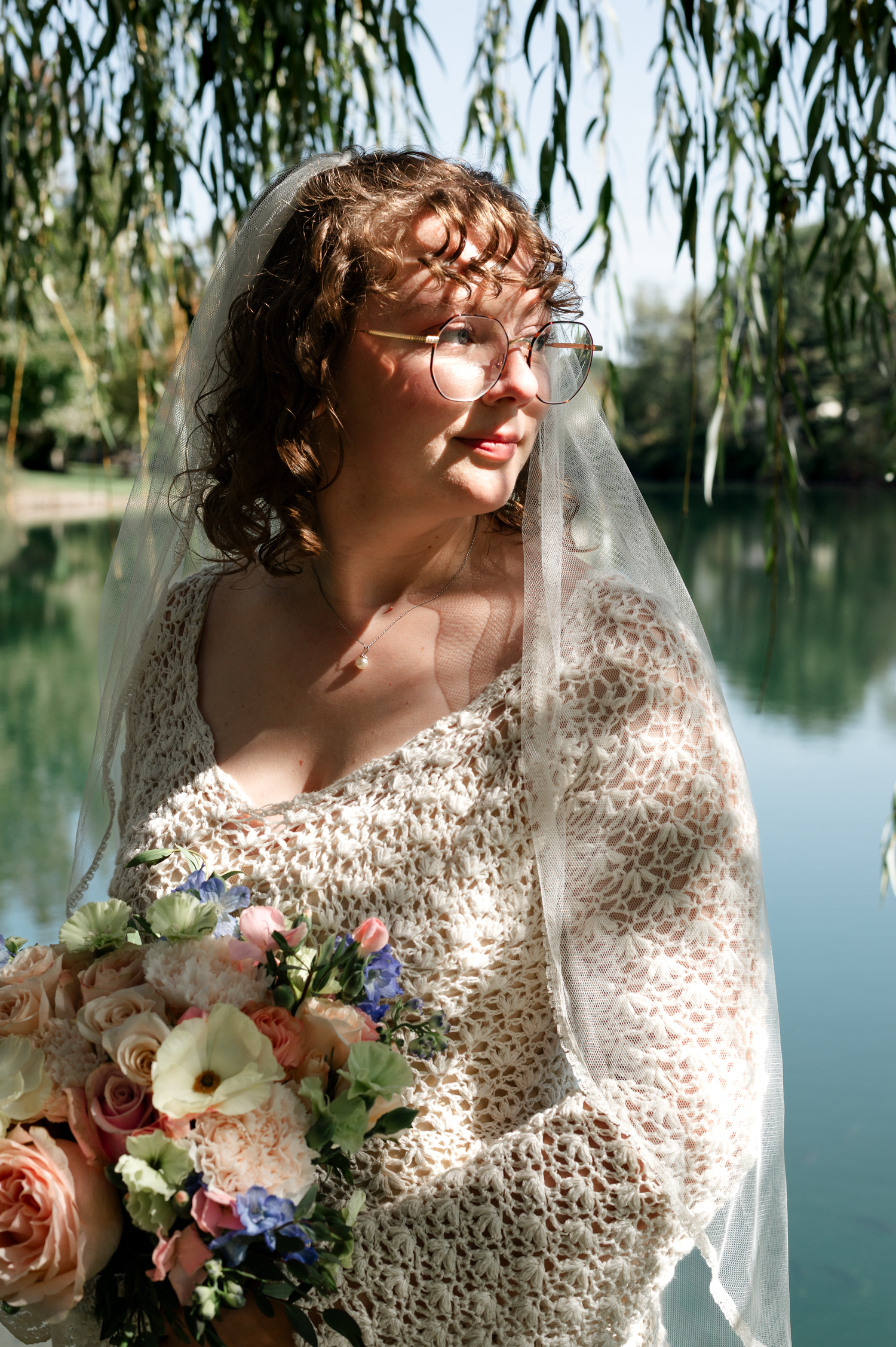 Bride in lace dress with veil holding bouquet by lakeside with willow trees.