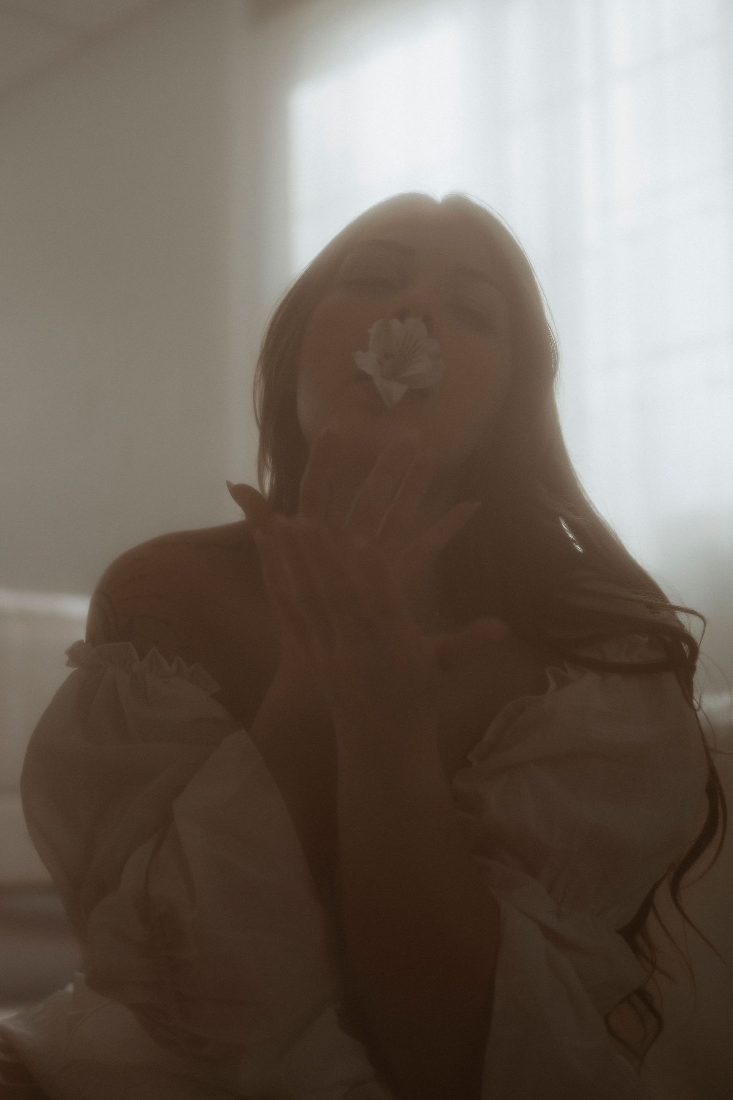 Woman in white off-shoulder top holding a white flower near her face in soft dreamy light.