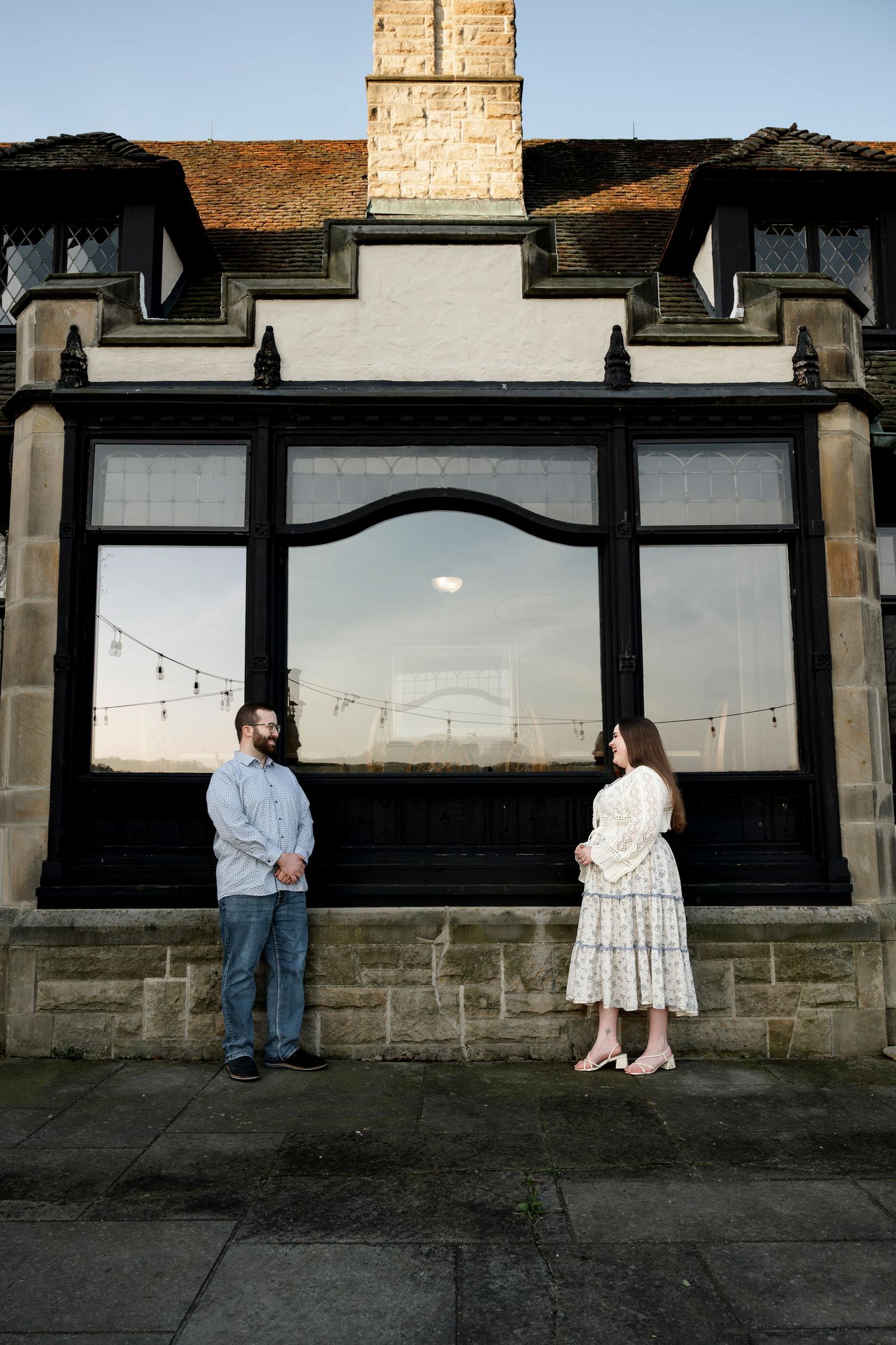 Couple standing apart before a vintage building with large black-framed windows reflecting string lights at dusk.