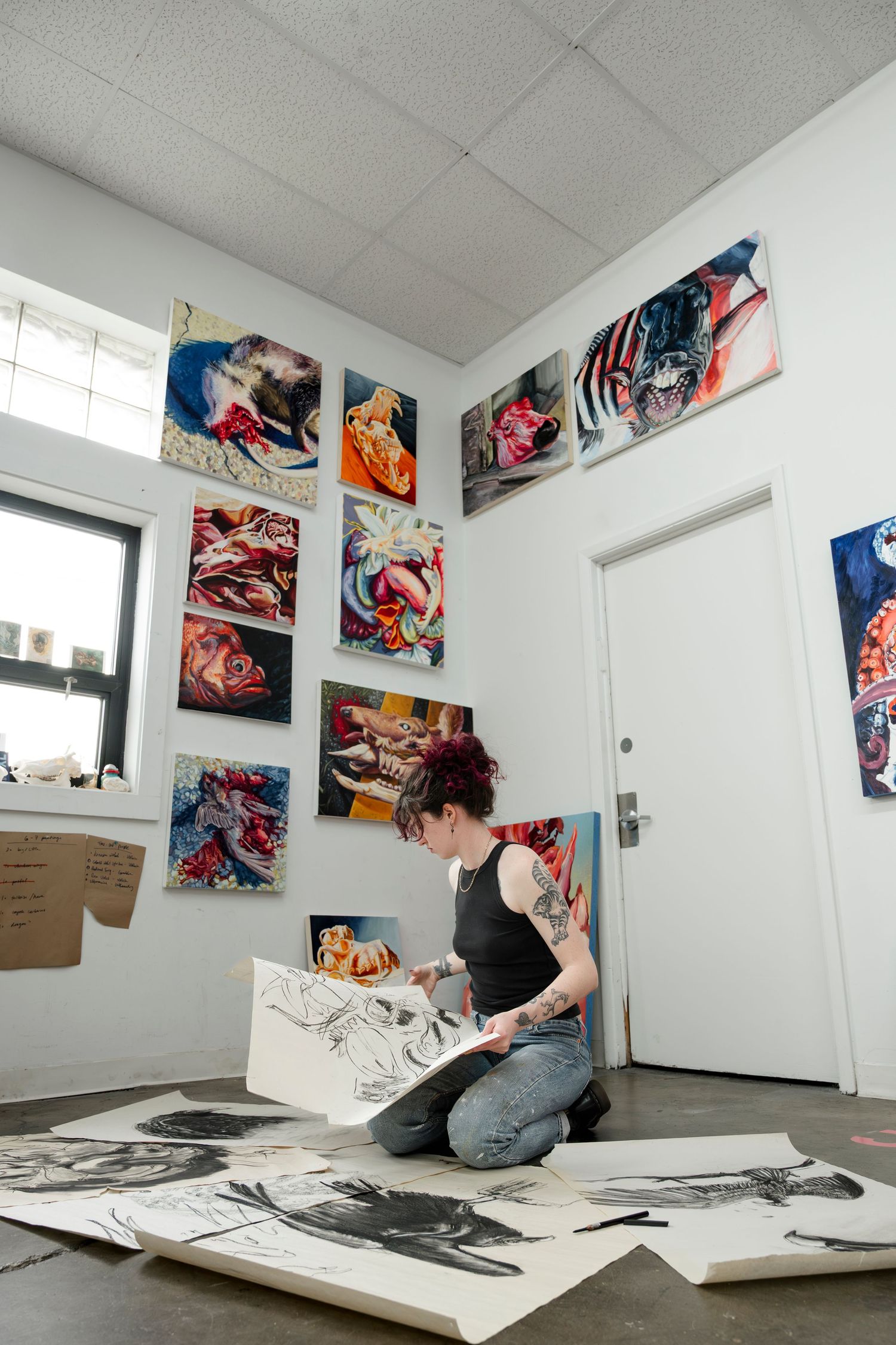 Artist working on floor sketches in studio space surrounded by vibrant abstract paintings on white walls.