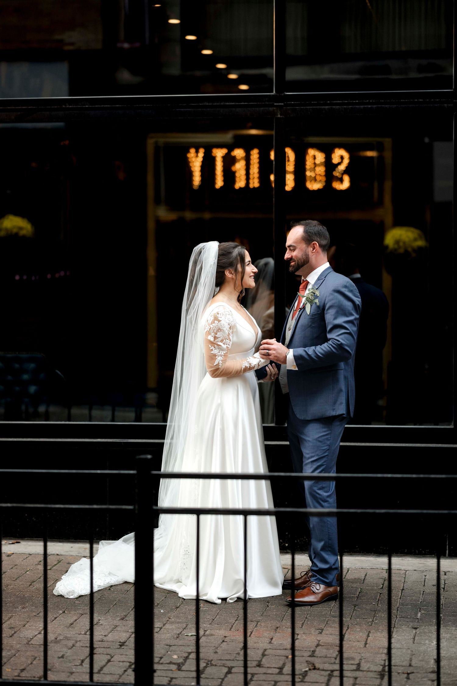 A romantic wedding couple shares a tender moment against a dark storefront background on a brick sidewalk.