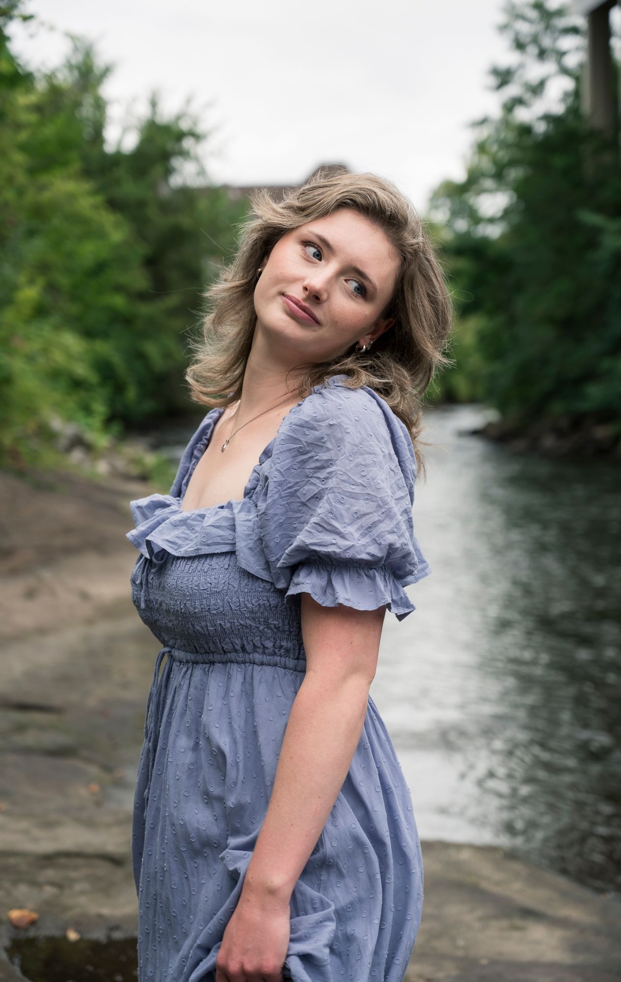 Person in blue ruffled dress poses by a flowing river on a summer day.