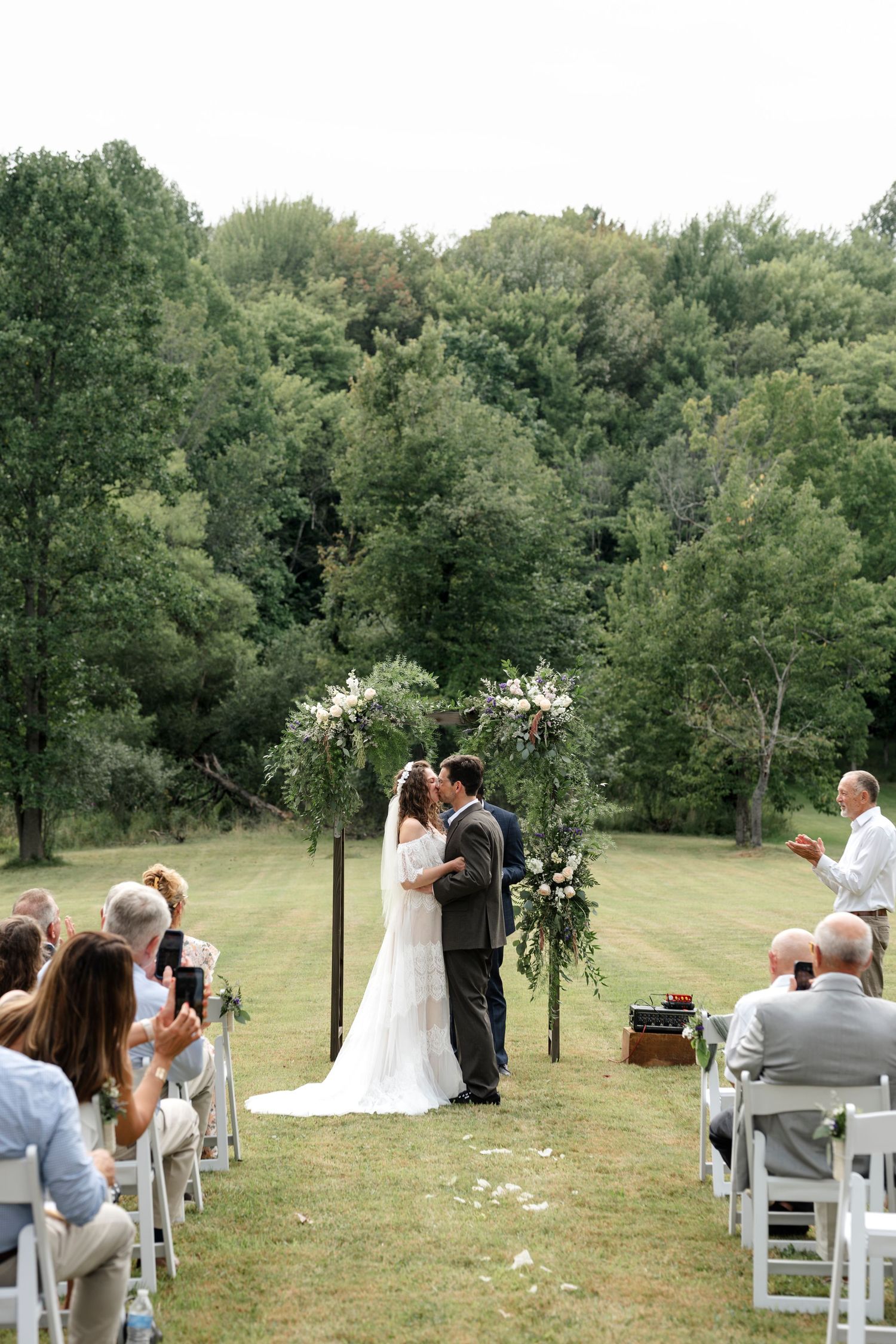 Bride and groom share first kiss at outdoor wedding ceremony surrounded by lush green trees.