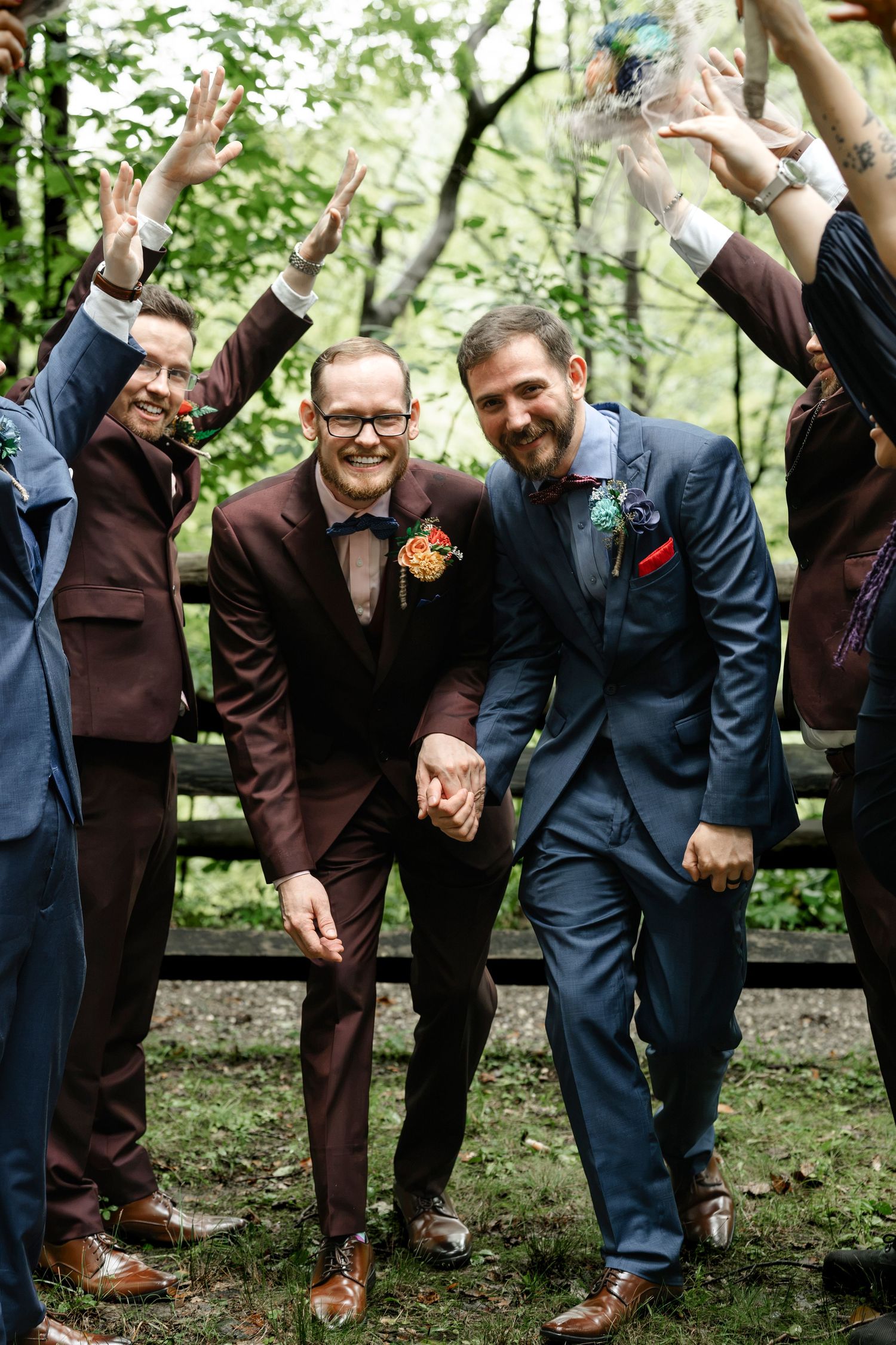 Two grooms hold hands and celebrate with wedding party in a lush green outdoor setting, wearing navy and burgundy suits.