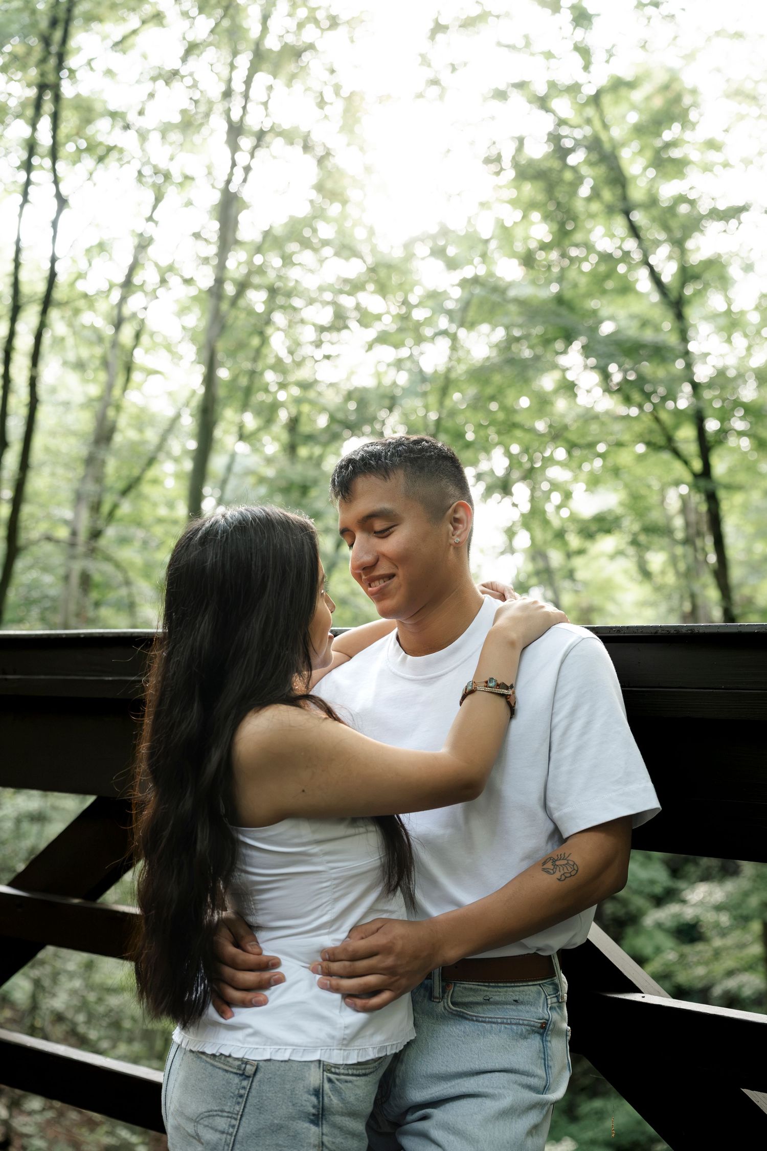 Young couple embracing on a wooden bridge surrounded by lush green forest trees in a romantic outdoor setting.