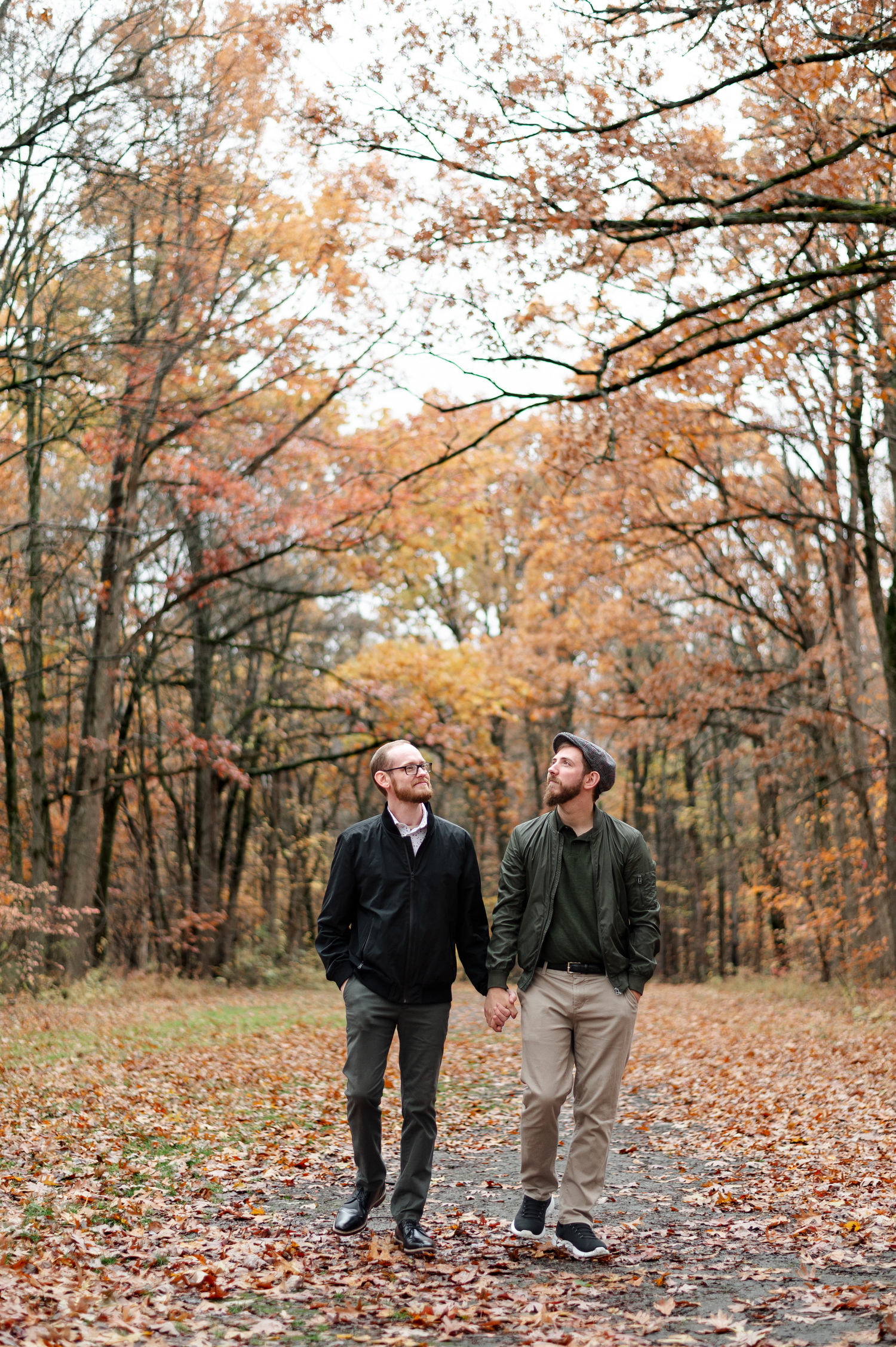 Two men holding hands in an autumn forest, surrounded by colorful fallen leaves and bare trees.