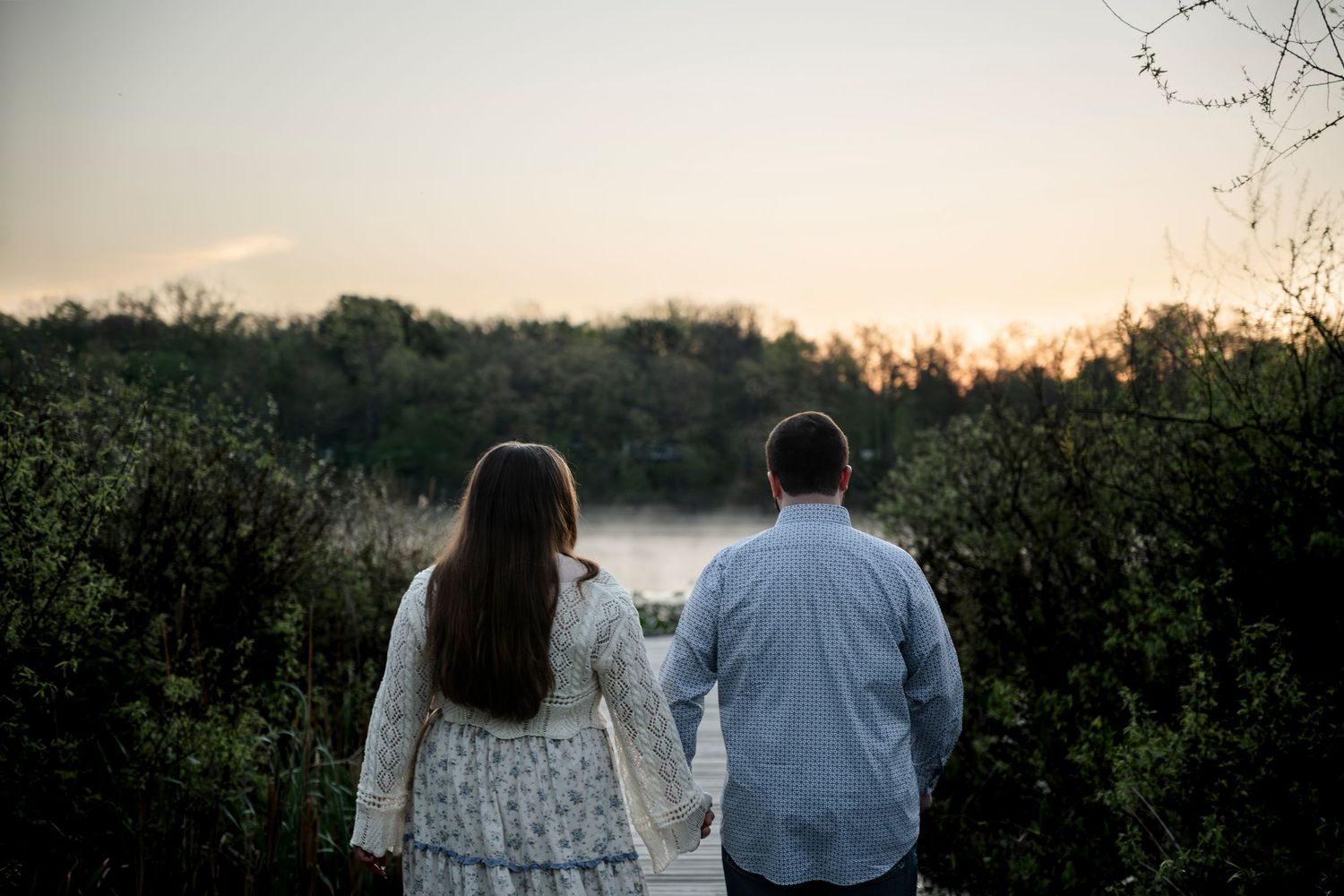 Couple holding hands facing a serene sunset over misty trees in a romantic outdoor engagement photo.