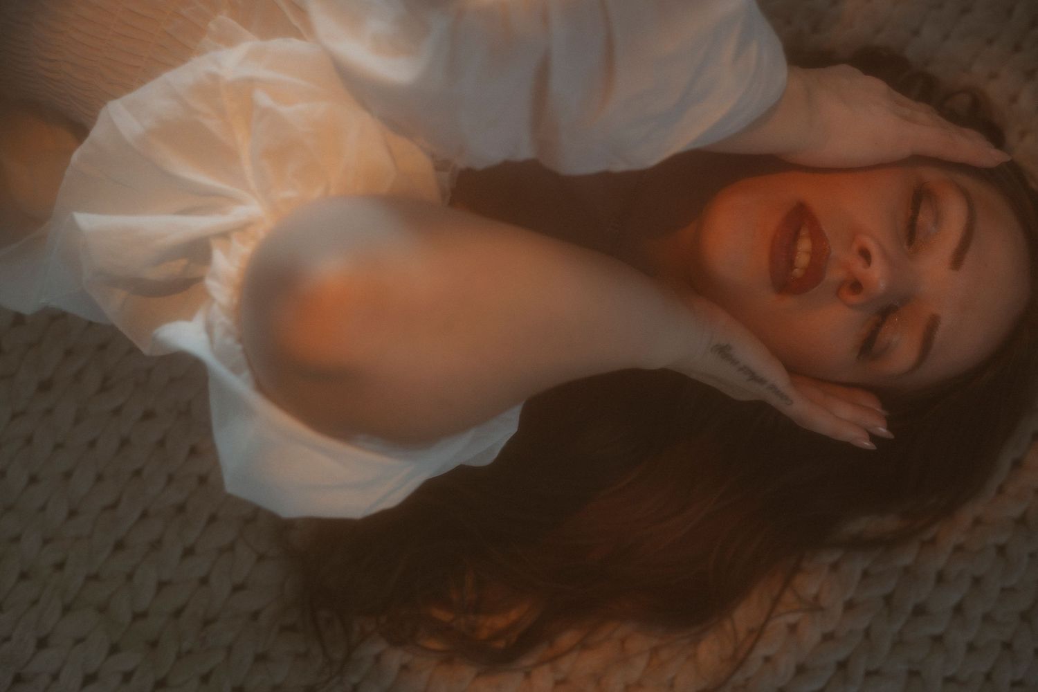 Young woman in white dress lying down, smiling with eyes closed in warm moody lighting.