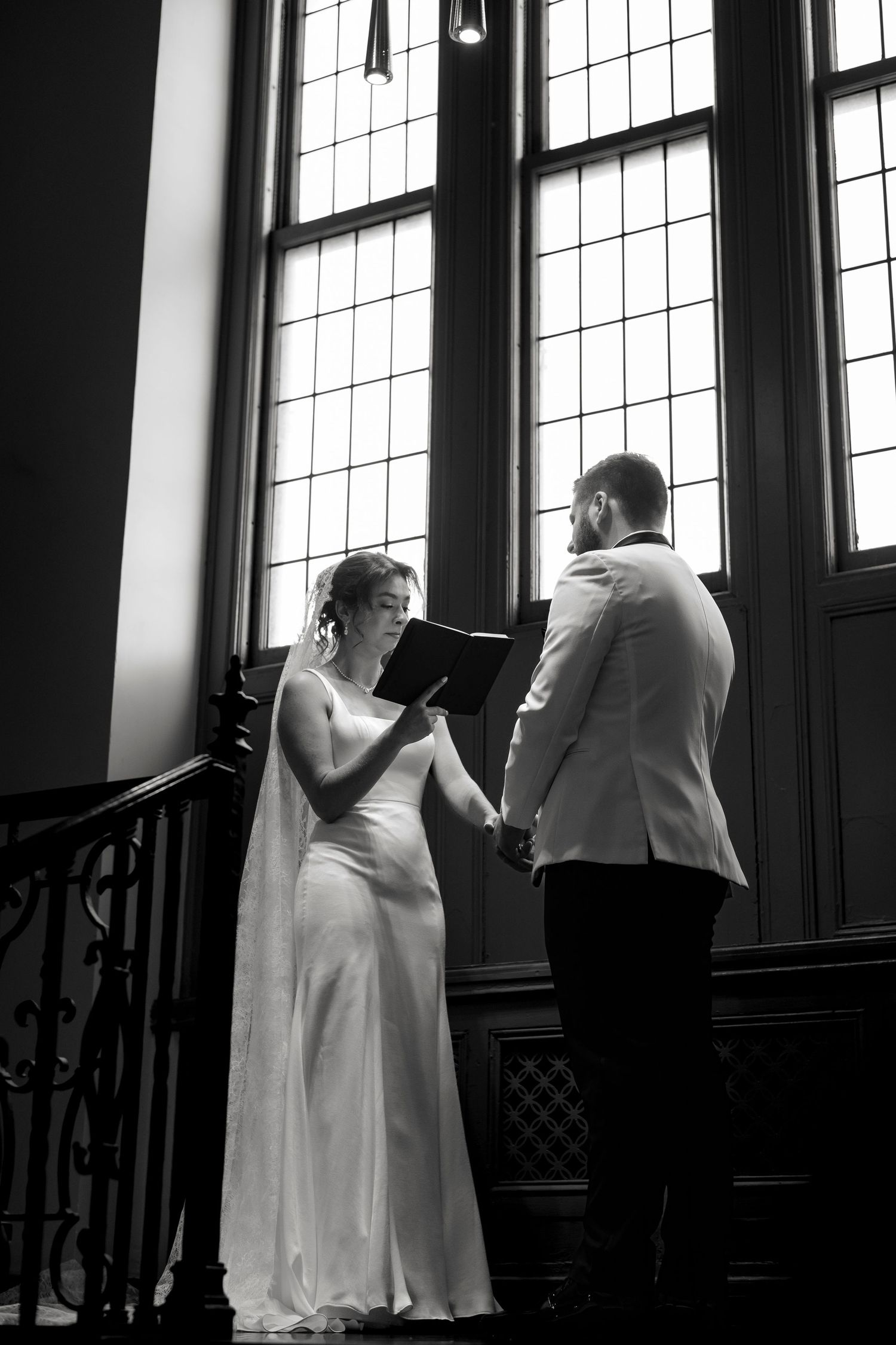 A black and white image of a bride and groom standing together in a formal setting with large windows in the background.